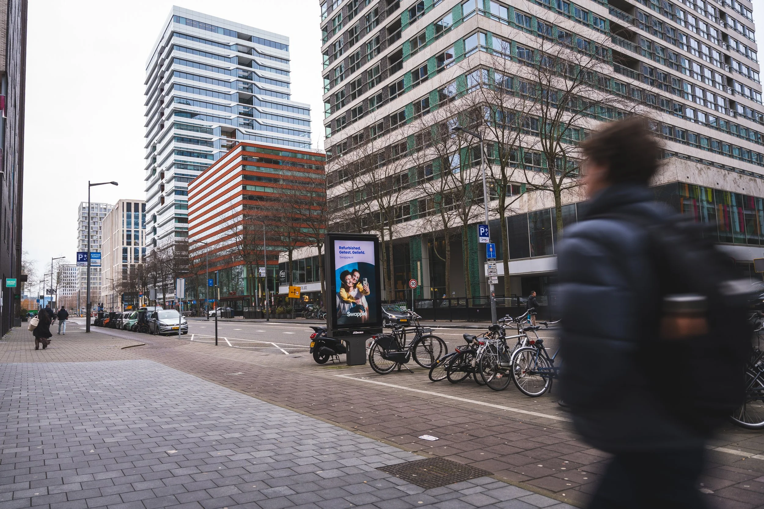 Stadstraat met hoge moderne gebouwen, fietsenstalling met fietsen, verkeersborden, en een bewegende persoon in de voorgrond.