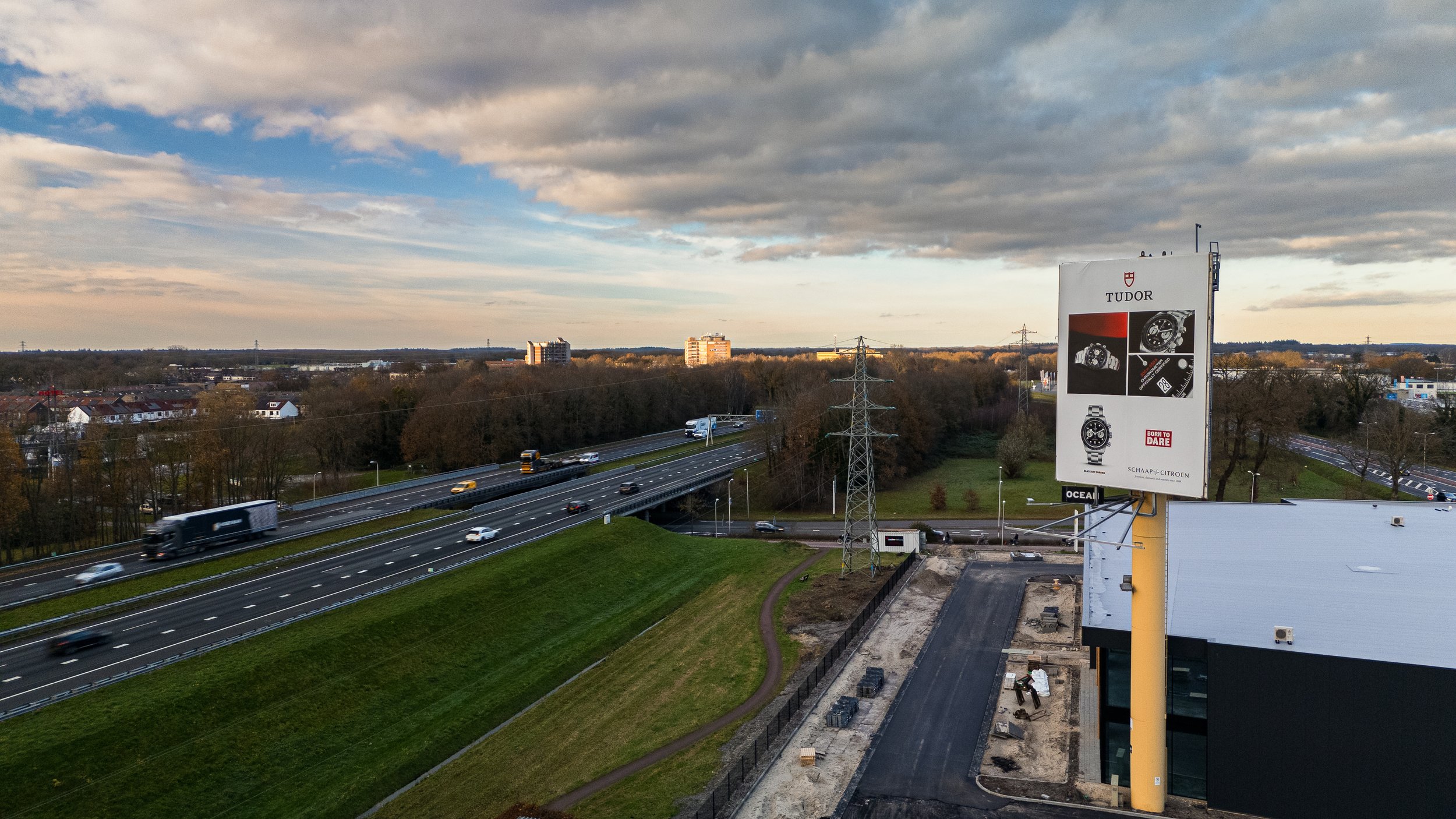 Uitzicht over een snelweg met verkeer, een groot advertentiebord met horloges en een gebouw onder constructie. Bos en woonwijken op de achtergrond onder een bewolkte hemel.