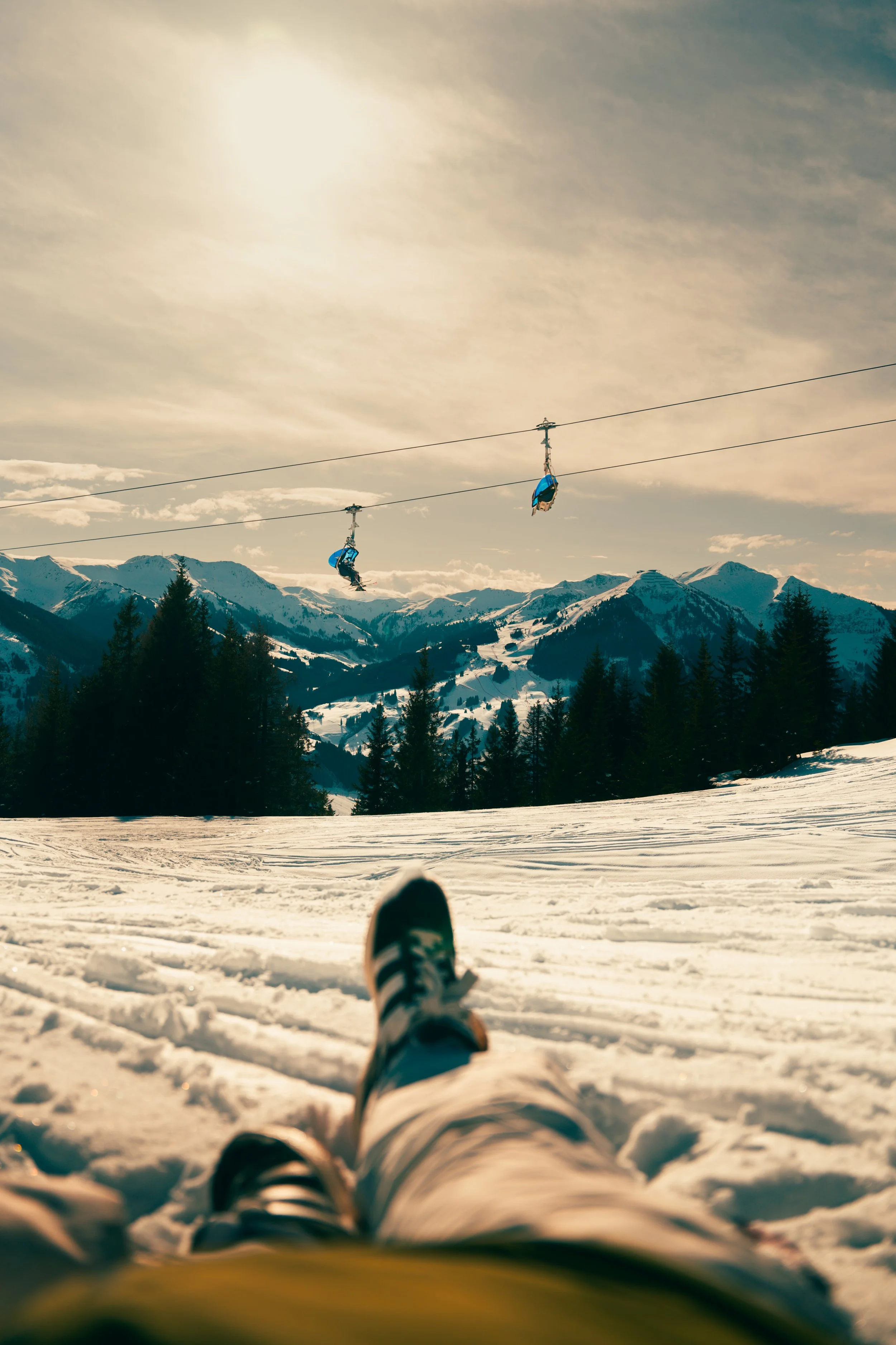 Persoon ligt op sneeuw met uitzicht op berglandschap en drie mensen op een kabelbaan in de lucht.