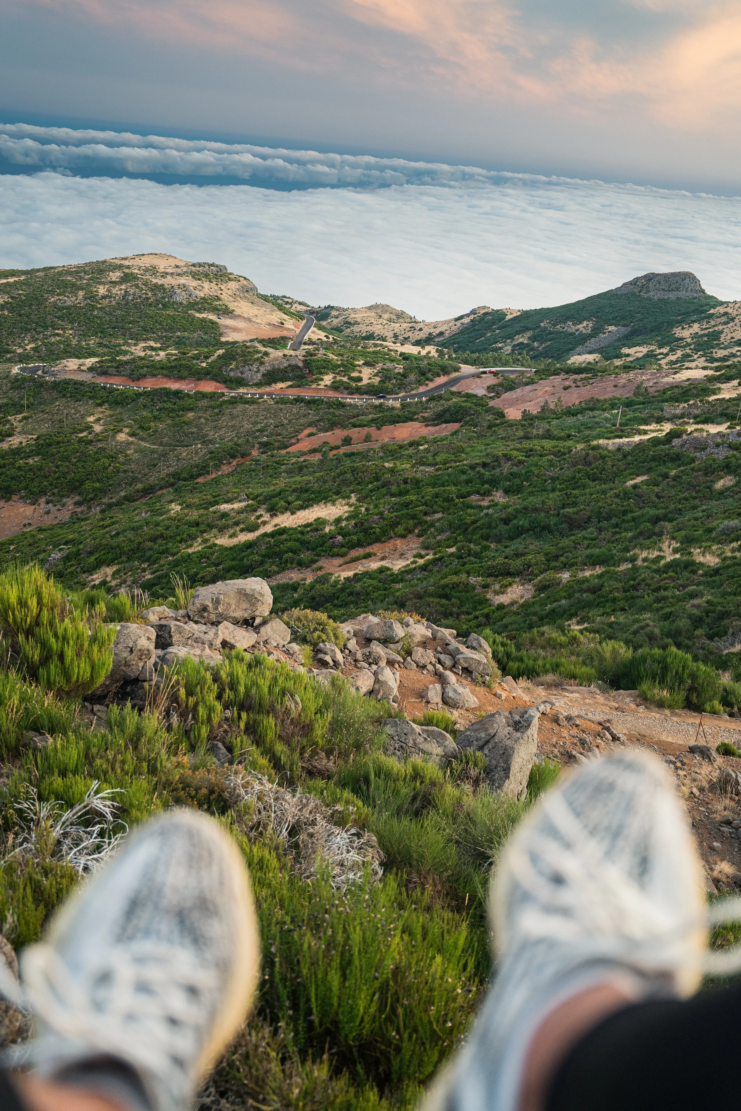 Perspectief vanaf de grond met witte sneakers, uitkijkend over een berglandschap met groene bossen, rotsen en een kronkelende weg onder een bewolkte hemel.