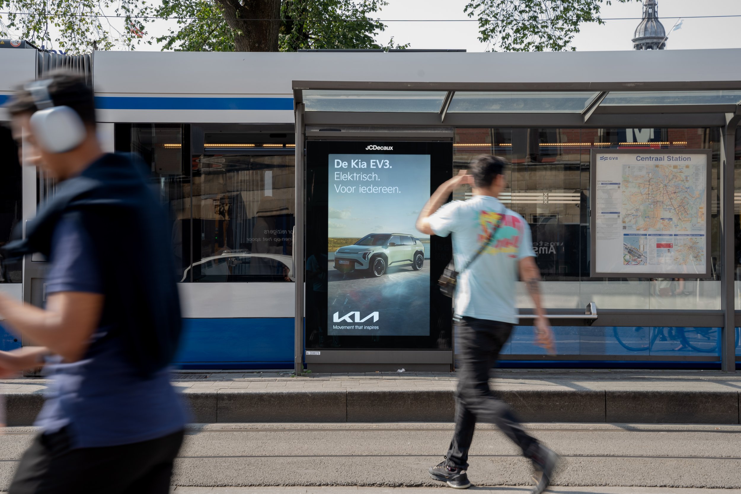 Mensen lopen langs een bushalte met een reclamebord voor de Kia EV3 elektrische auto, naast een stadsplattegrond en een bus. Er rijdt een blauwe tram op de achtergrond.