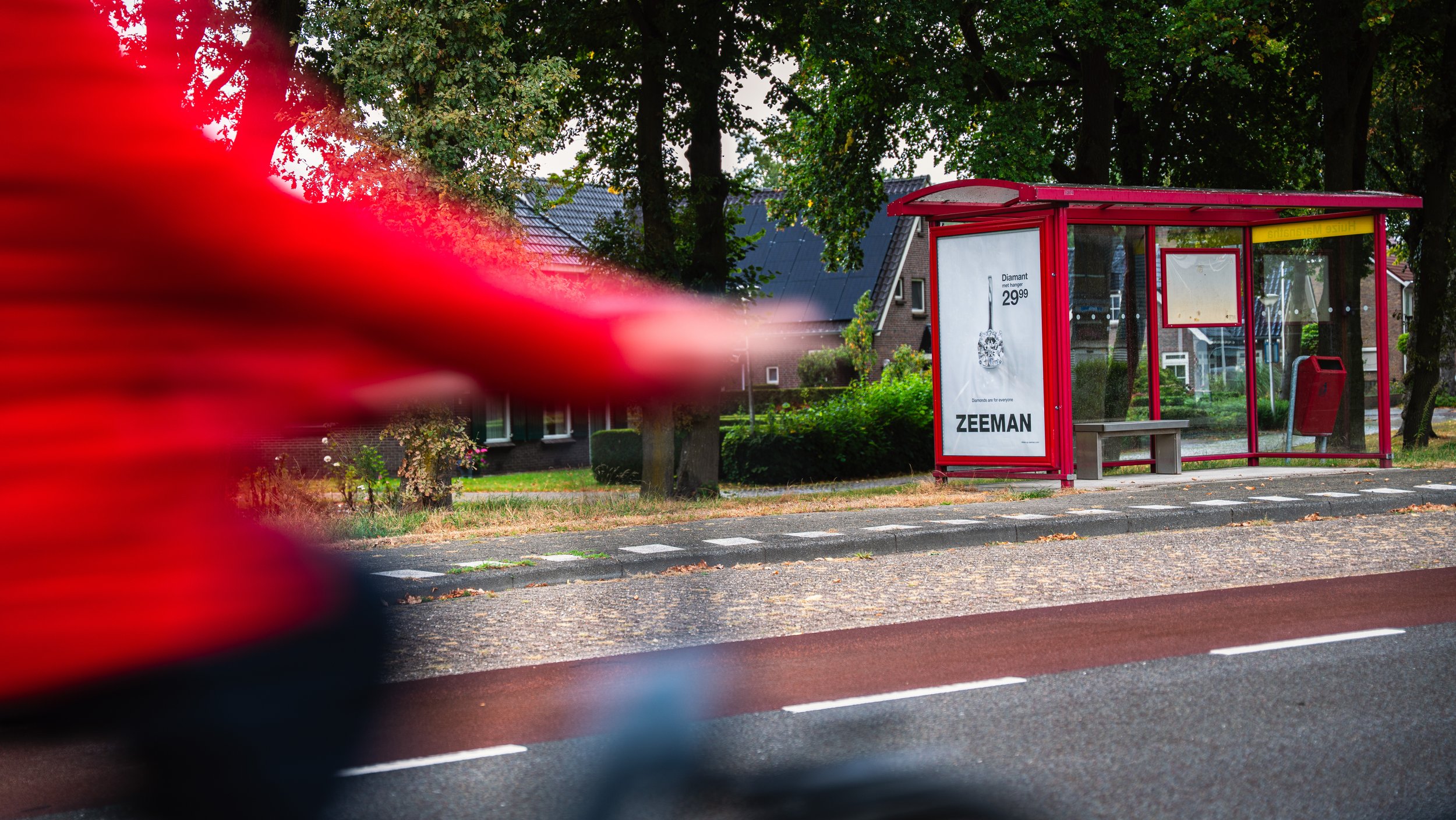 Een bushalte met een rood metalen frame en een advertentiebord, omringd door groene bomen en woningen op de achtergrond, met een rode fietser op de voorgrond.