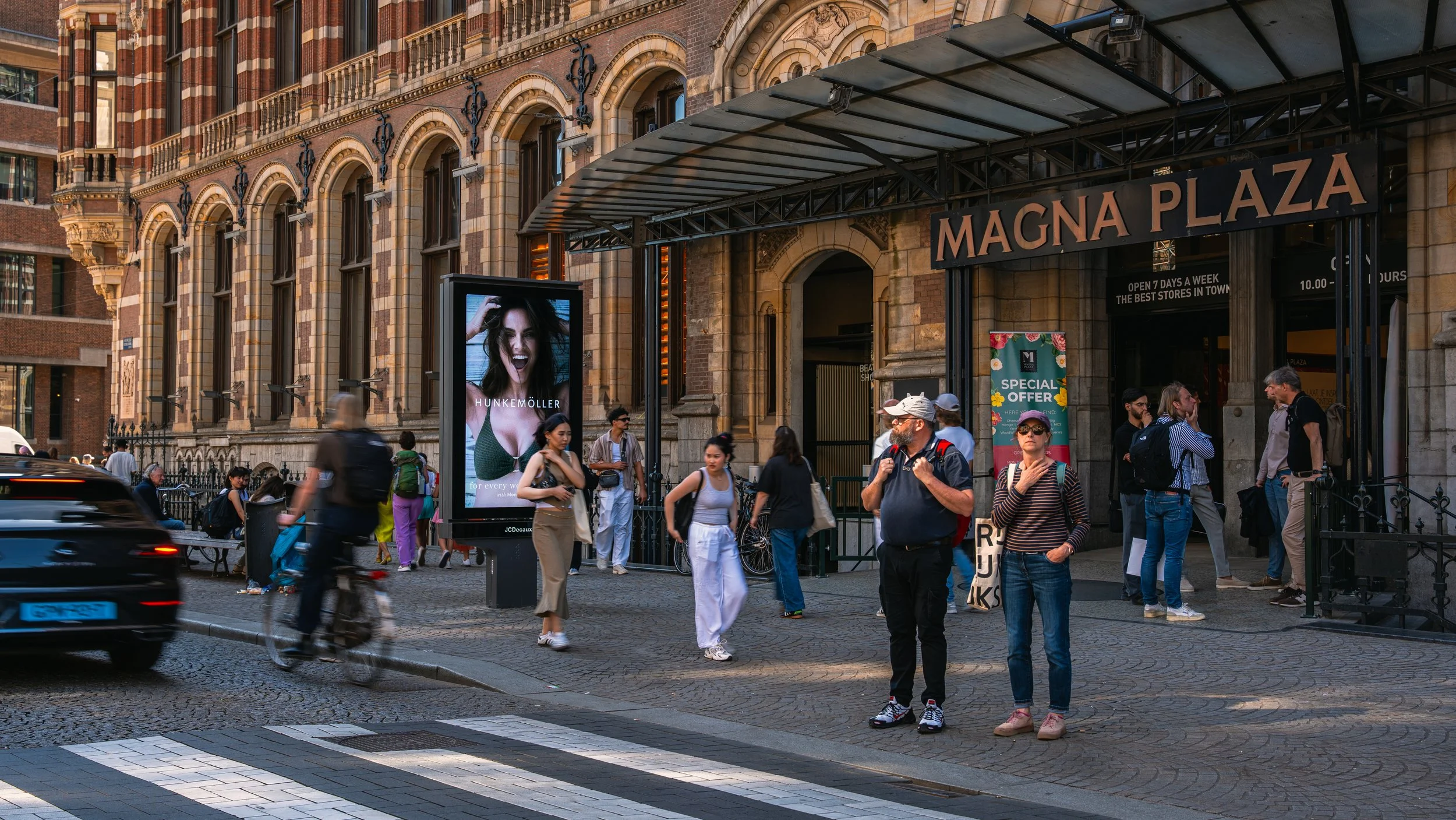 Straatbeeld voor de ingang van Magna Plaza met mensen die wachten en passeren, een digitaal reclamebord en een bord met 'Special Offer'.