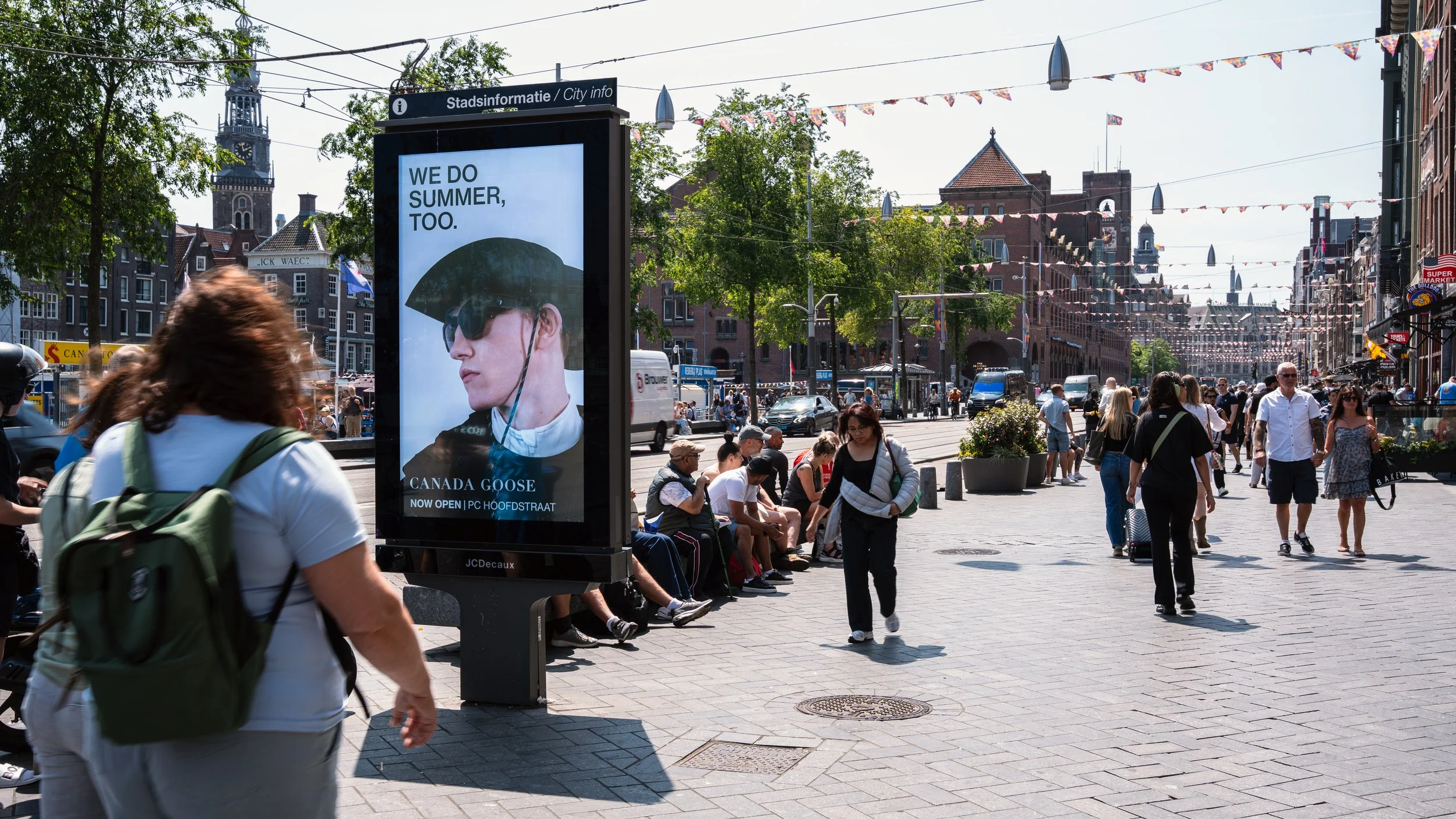 Voorbeeld van een drukke stadsstraat met mensen die lopen en wachten op banken, een reclamebord met een afbeelding van een persoon met hoed en donkere brillen, en historische gebouwen op de achtergrond.