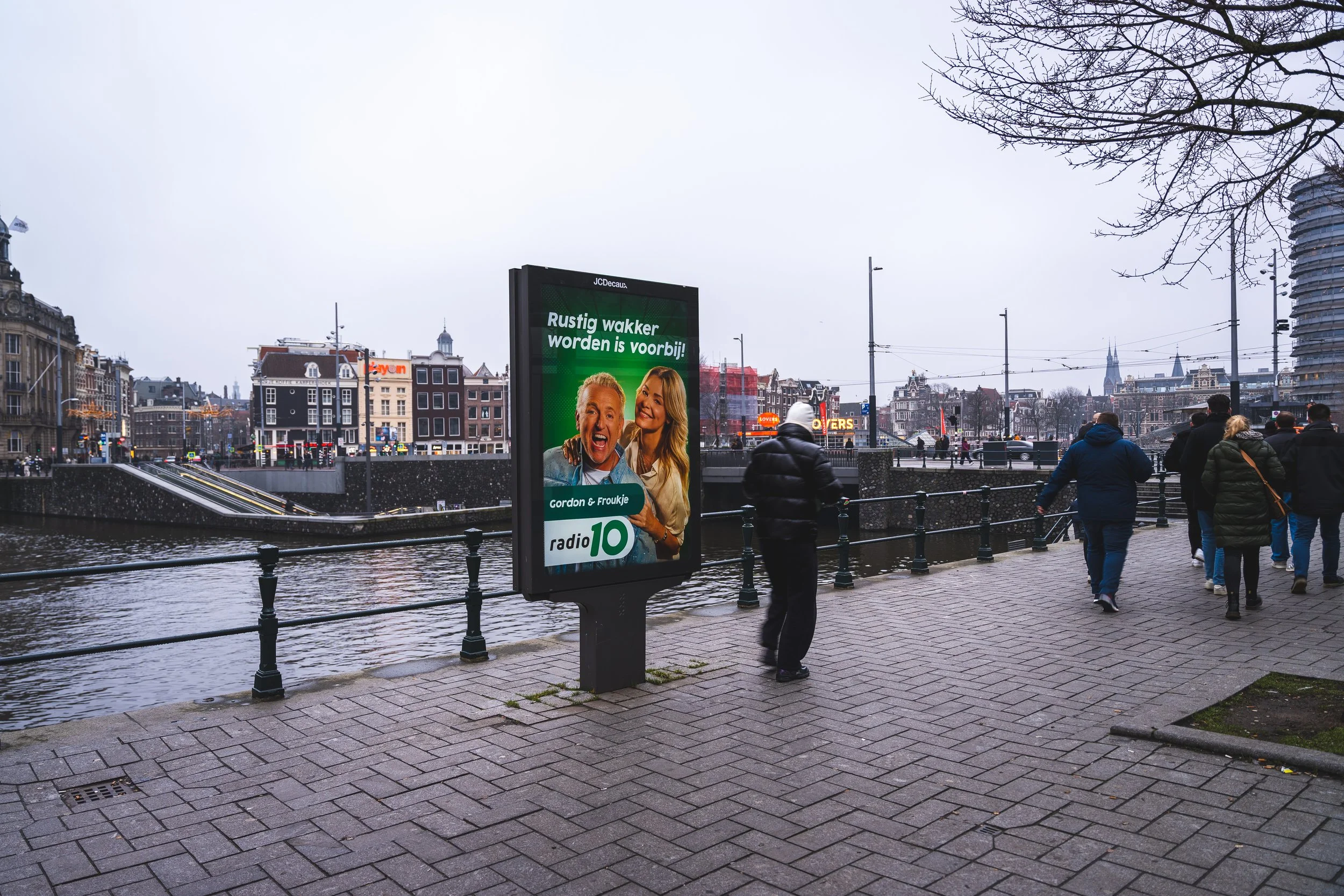 Straatbeeld in Amsterdam met mensen die wandelen langs de gracht en een digitaal reclamebord met een advertentie voor radio 10, tegen een grijze, bewolkte hemel.