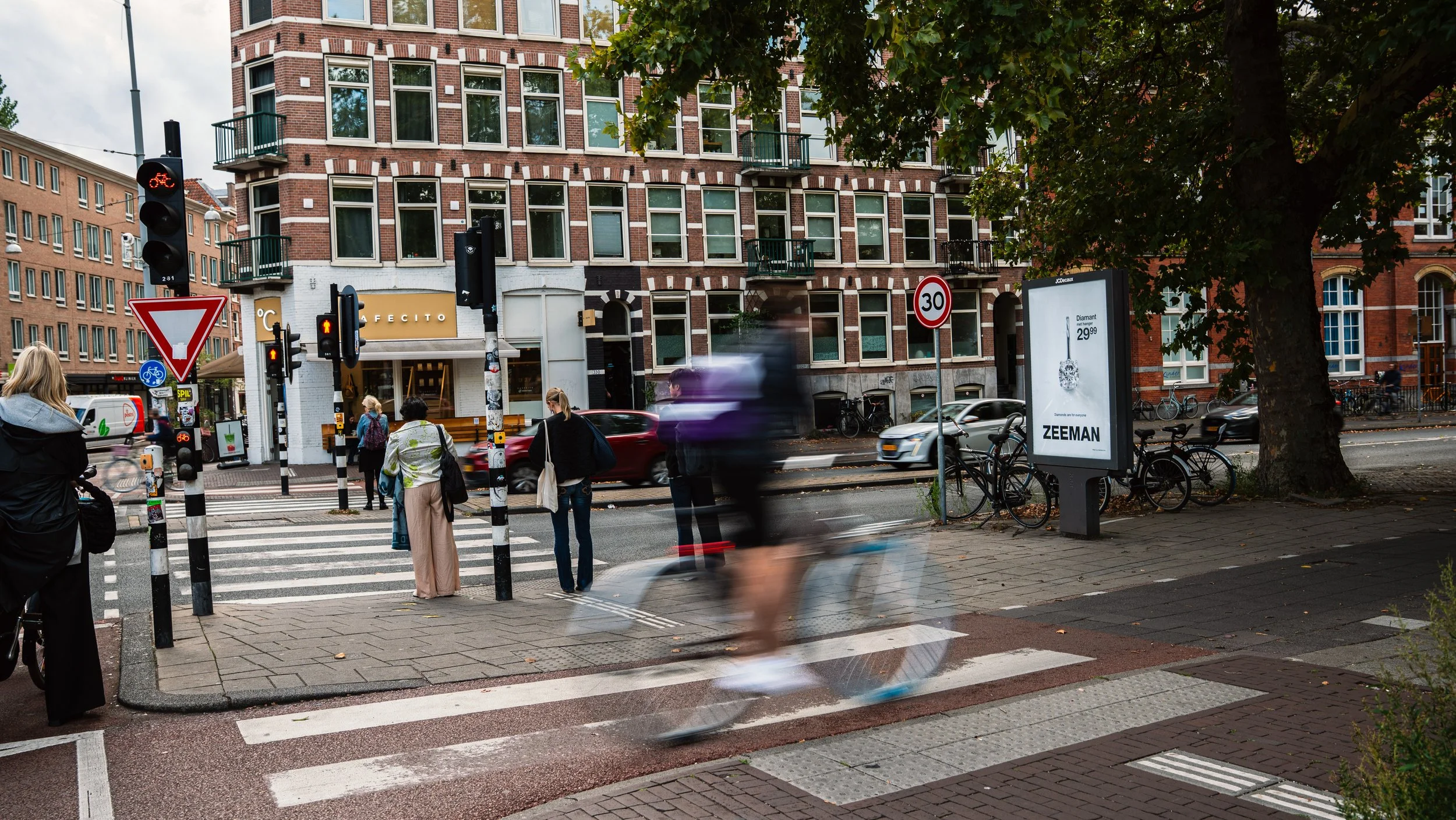 Straatbeeld met voetgangers en fietsers, verkeerlichten en een grote boom op de hoek van de straat, in een stedelijke omgeving met historische gebouwen en reclameborden.