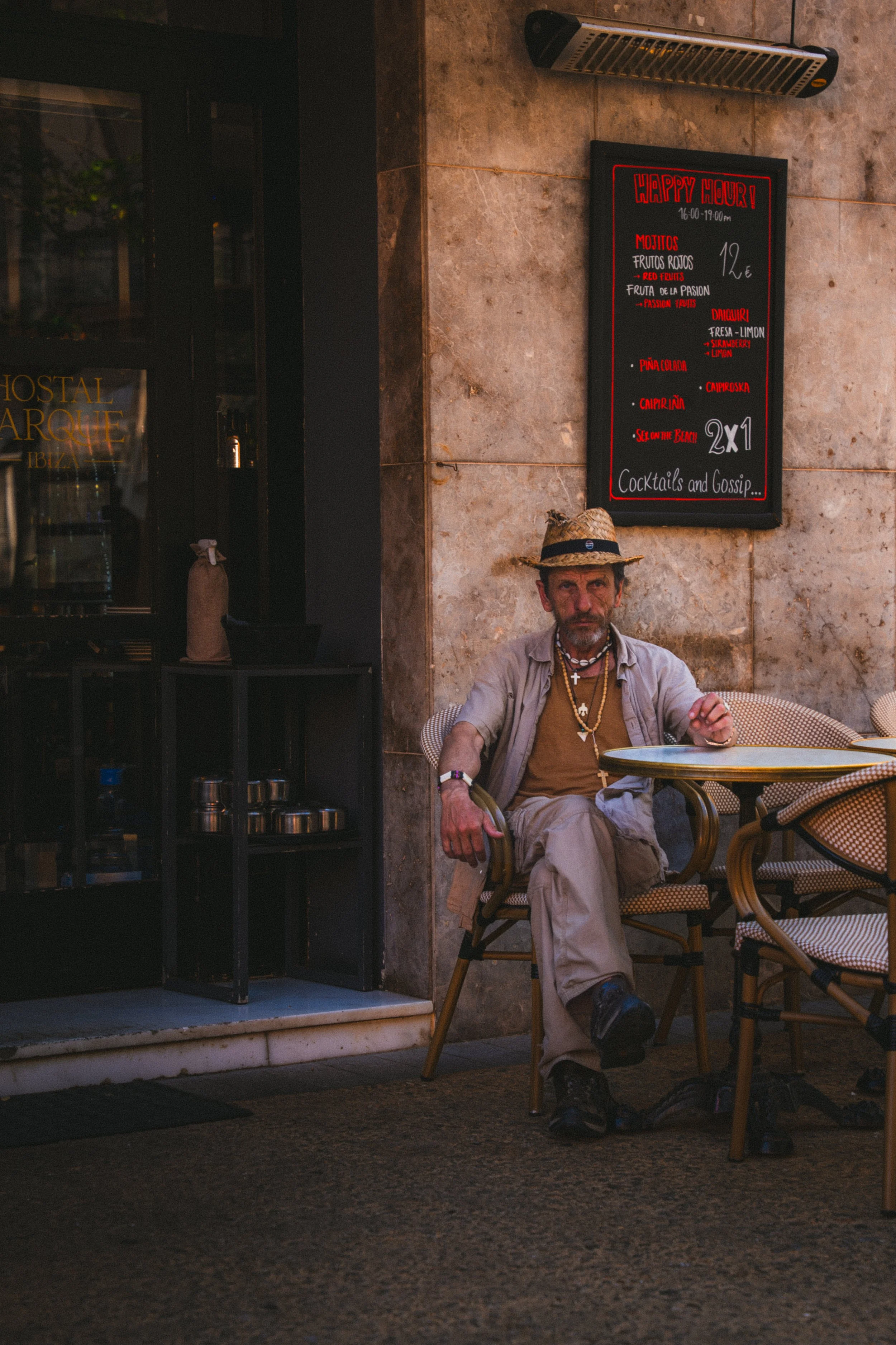 Man zit buiten op een terras, draagt hoed, sieraden en casual kleding, naast een tafel. Op de muur hangt een bord met een happy hour aanbieding op cocktails.