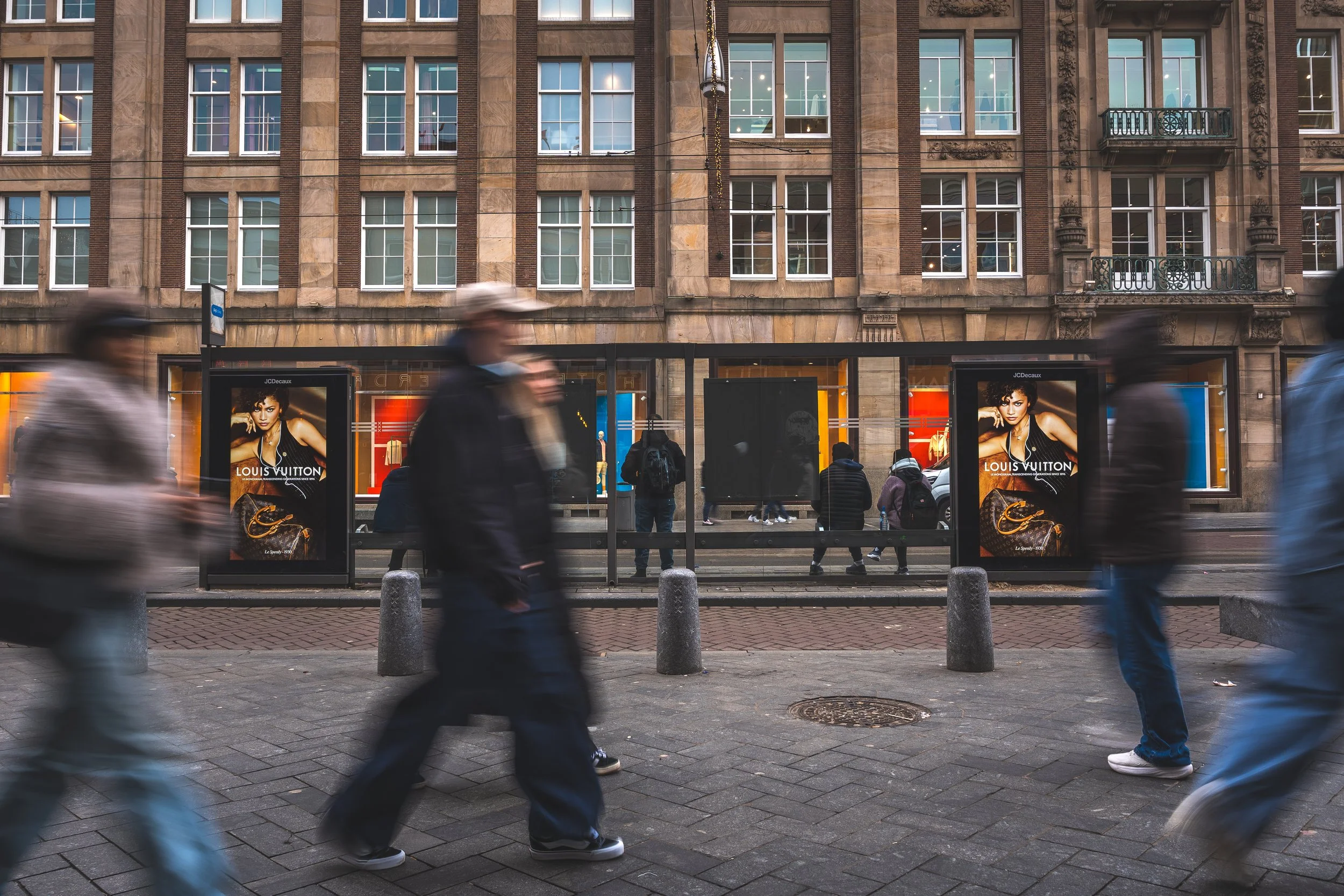 Straatbeeld met lopende mensen voor een bushokje en een winkelpand met Louis Vuitton advertenties.