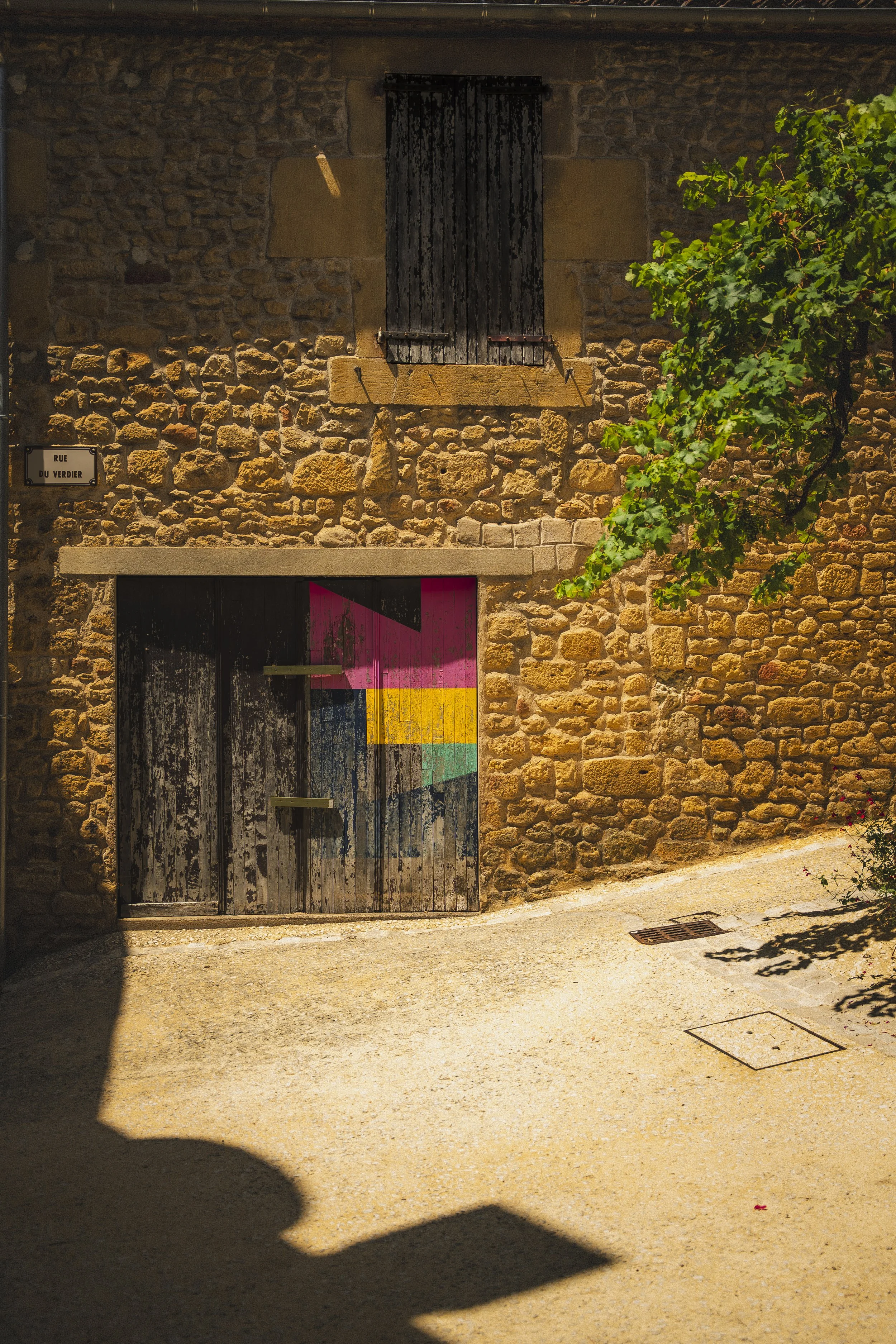 Een stenen muur met een dichtgeklapt zwart houten raam en een grote houten deur met kleurrijke geometrische patronen. Een groene klimop groeit langs de muur, en er is een straatbord dat 'Rue du Verdier' aangeeft.