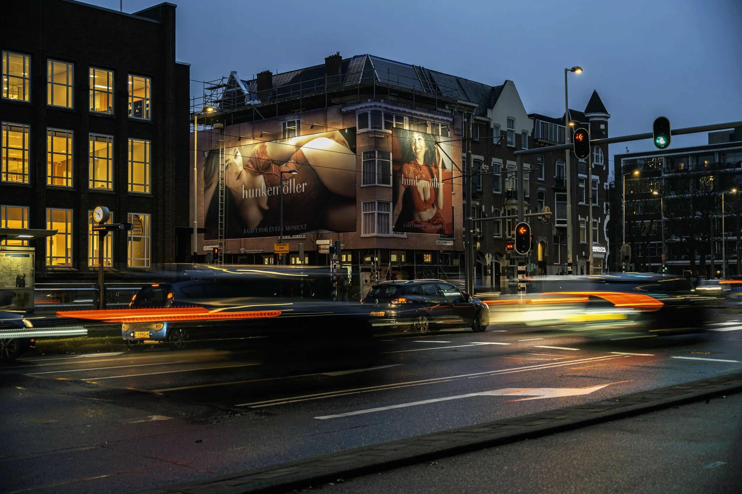 Een stedelijk straatbeeld tijdens de avond met bewegende auto's en verlichte gebouwen, inclusief grote advertenties op een gebouw.