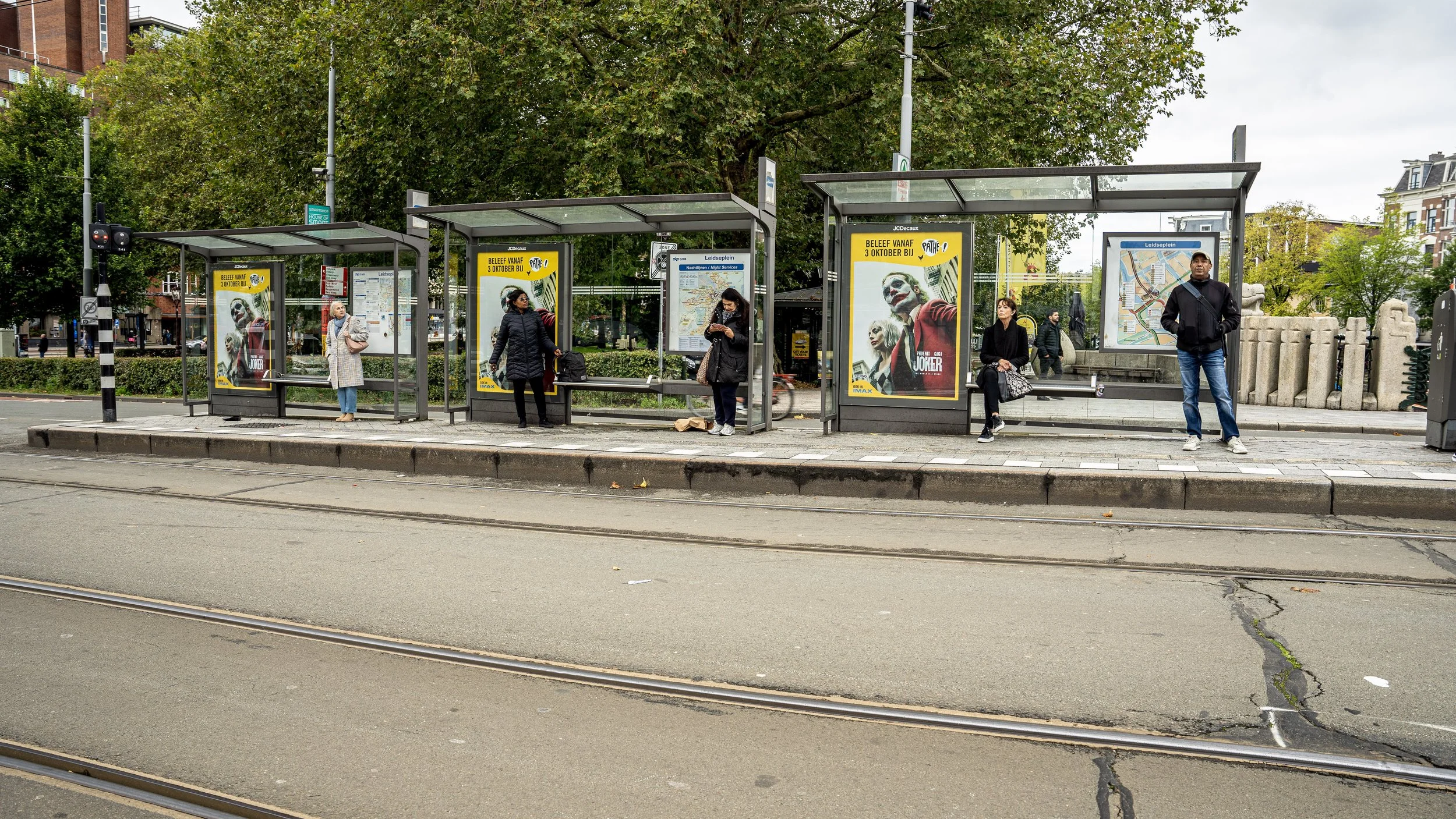 Mensen wachten bij een bushalte in een stad met tramrails op een straat. Er zijn vier mensen te zien, sommigen zitten en anderen staan. Verschillende posters en een stadsplan hangen in de bushalte. Er is groen groen in de achtergrond en gebouwen op d