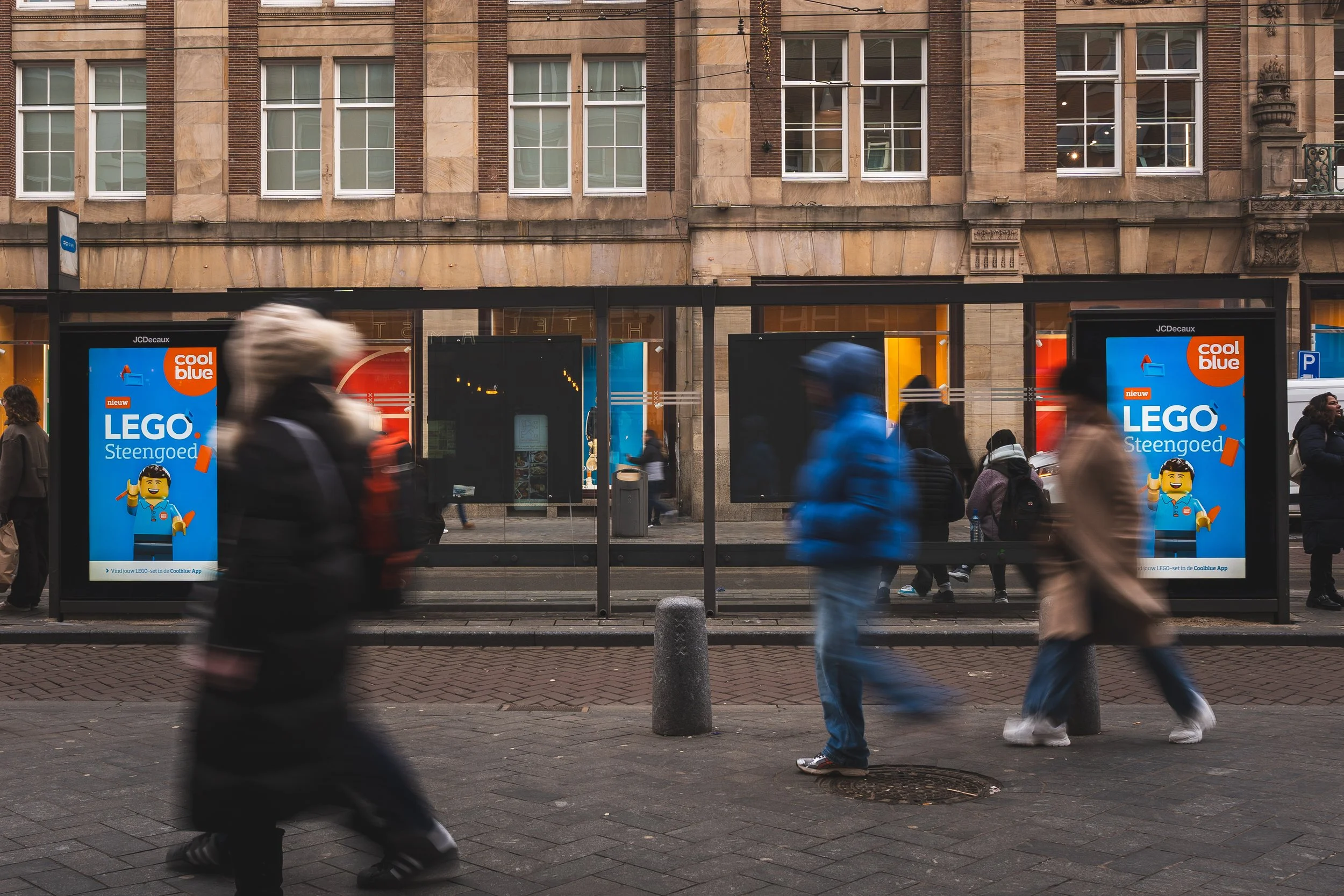Bezoekers wachten bij een bushokje op een drukke stedelijke halte, met advertenties met LEGO en Coolblue. Mensen lopen voorbij, sommige bewegen snel, others staan stil.