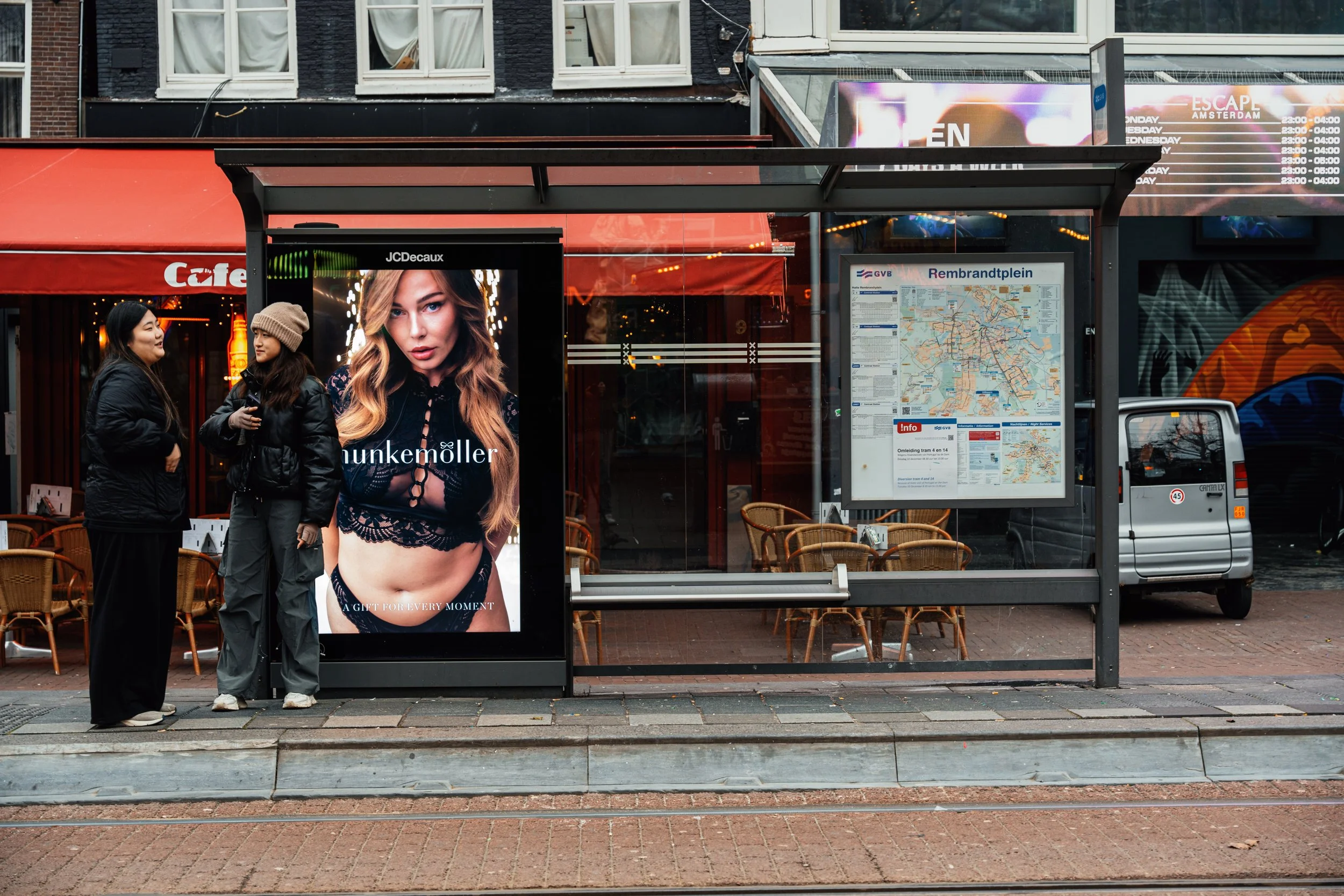 Twee vrouwen spreken elkaar bij een bushokje in Amsterdam, met een grote reclameposter en een plattegrond van de stad op de achtergrond.