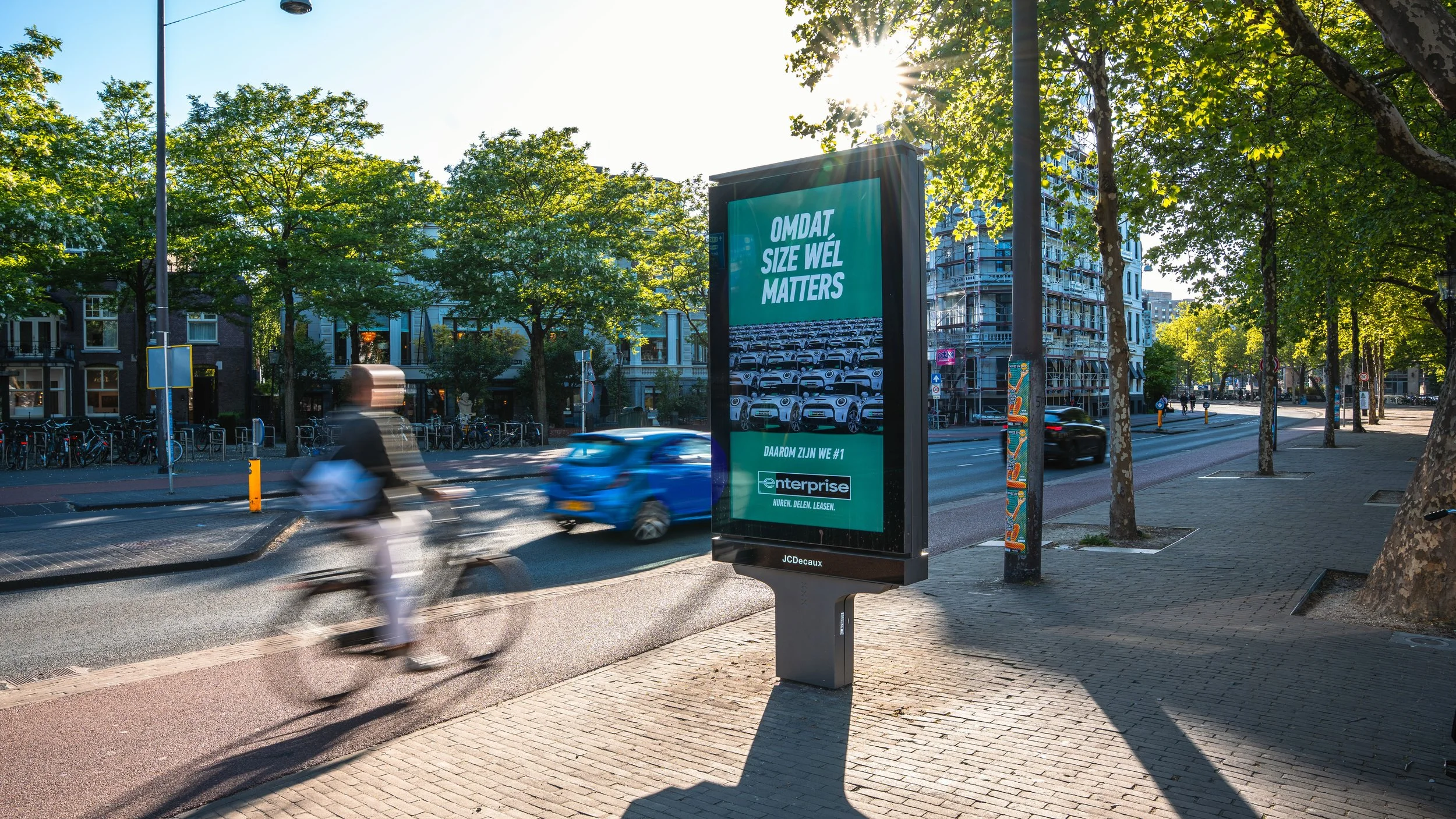 Elektronisch reclamebord op straat met tekst 'Omdat size wél matters' en een afbeelding van meerdere auto's, met bomen en gebouwen op de achtergrond, en een fietser die voorbij rijdt.