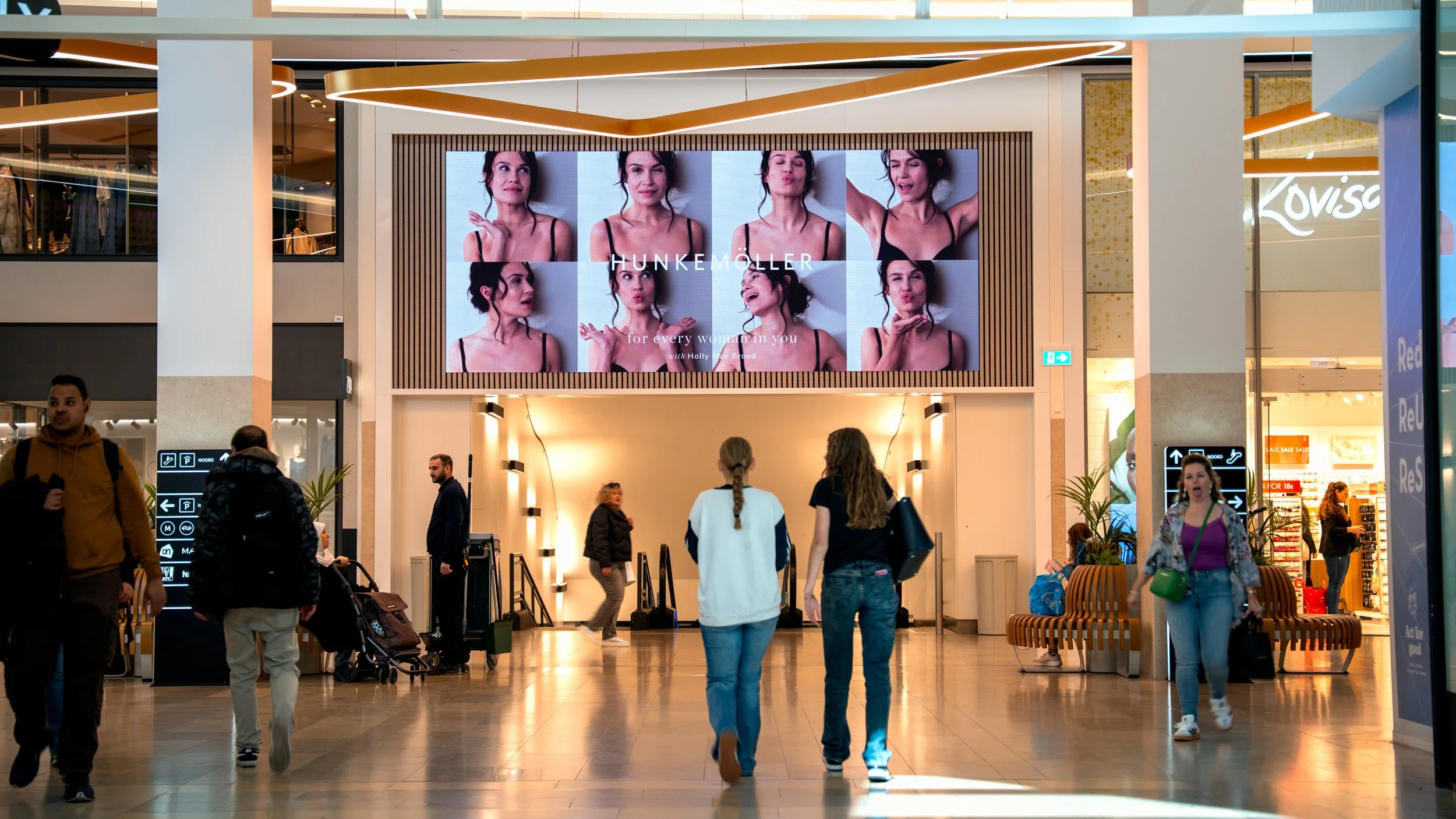 Binnenkomst in een winkelcentrum met grote digitale schermen, drie mensen lopen naar binnen en er zitten enkele mensen op banken.