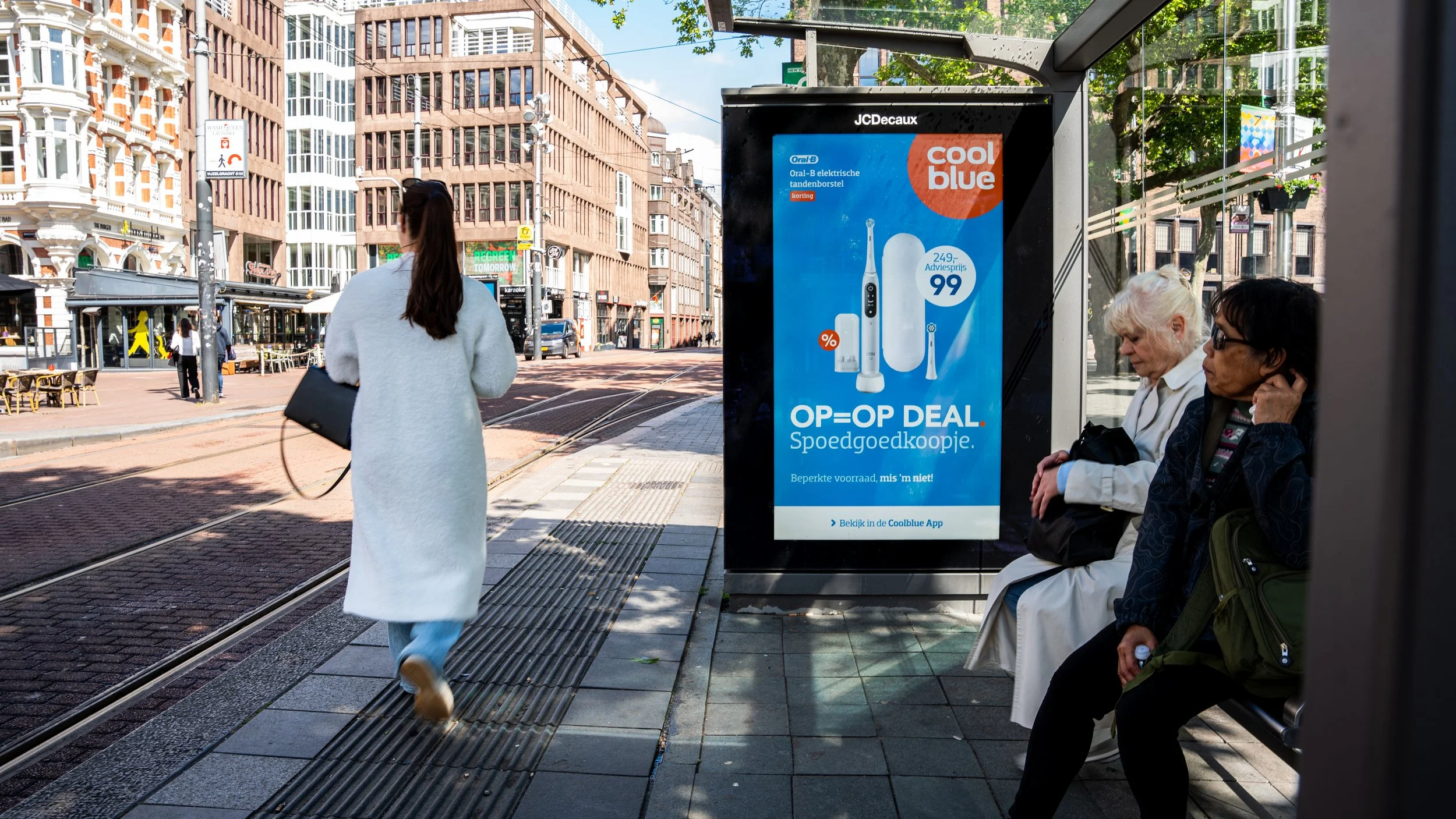 Straatbeeld met een vrouw die wegloopt, een advertentie van een elektrische tandenborstel en twee vrouwen zittend op een bankje.