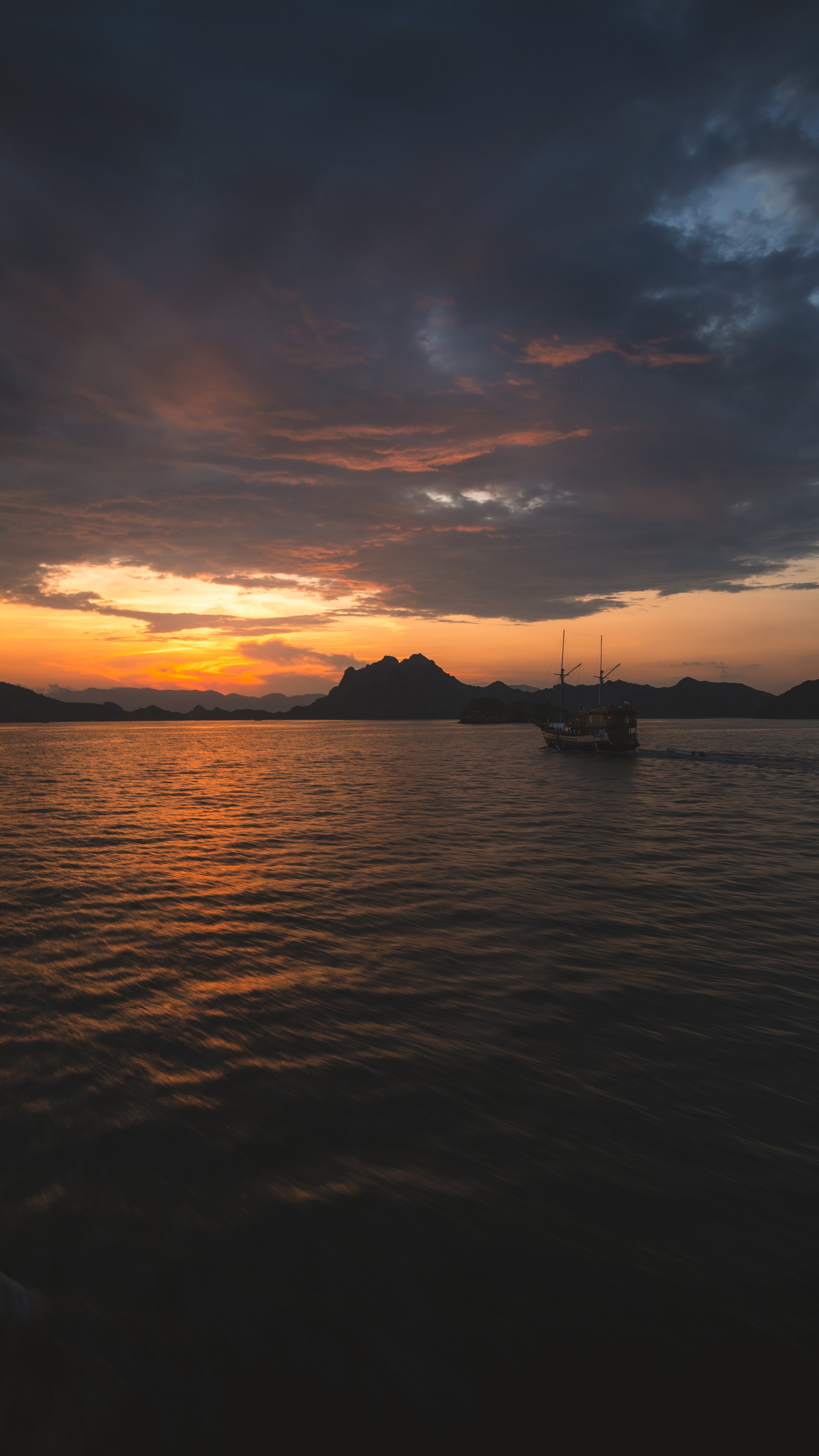 Een boot op rustig water tijdens zonsondergang met donkere wolken en bergen op de achtergrond.