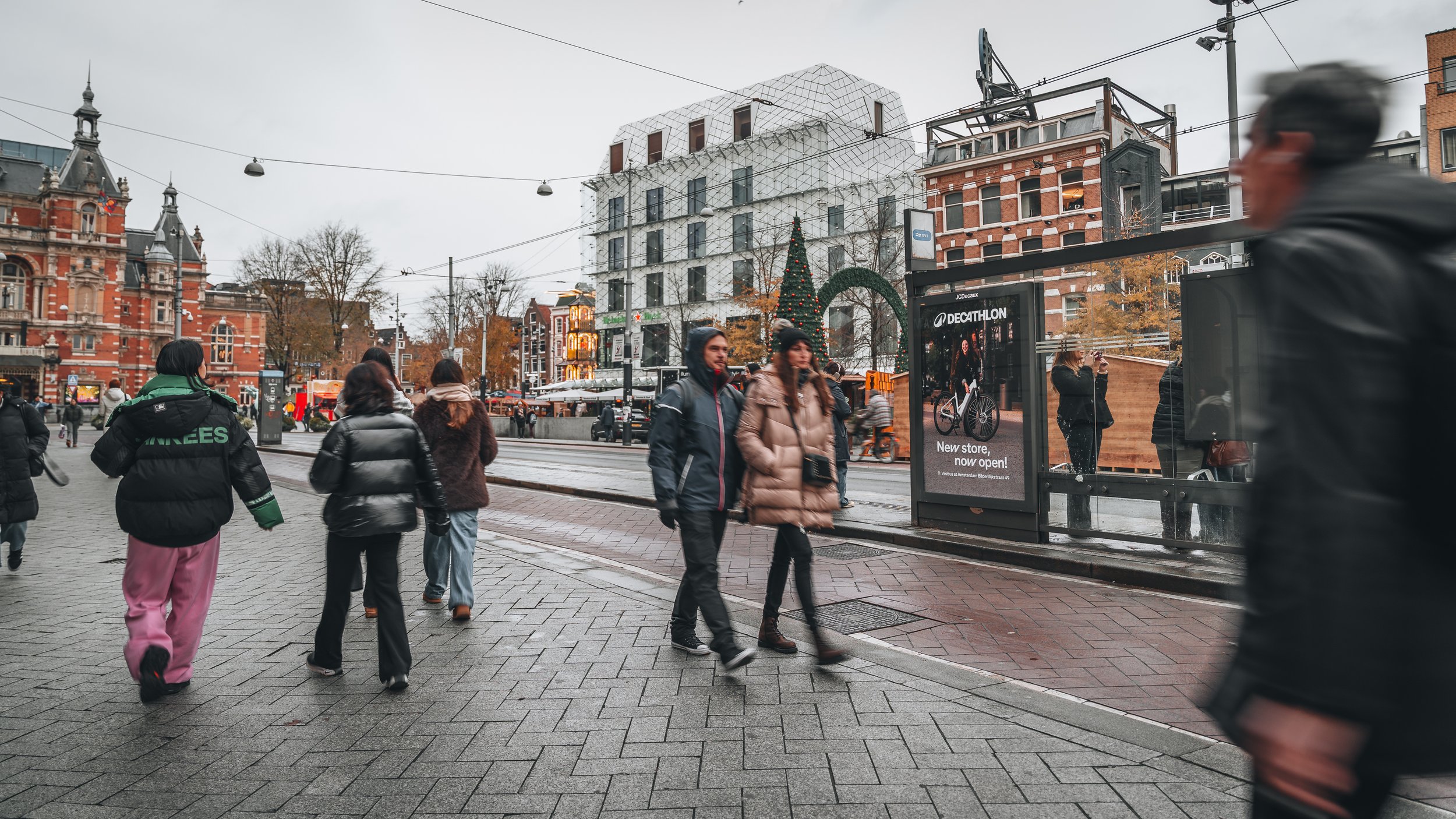 Stadsscene met mensen op een tramhalte, een versierde kerstboom en historische gebouwen op een grijze dag.