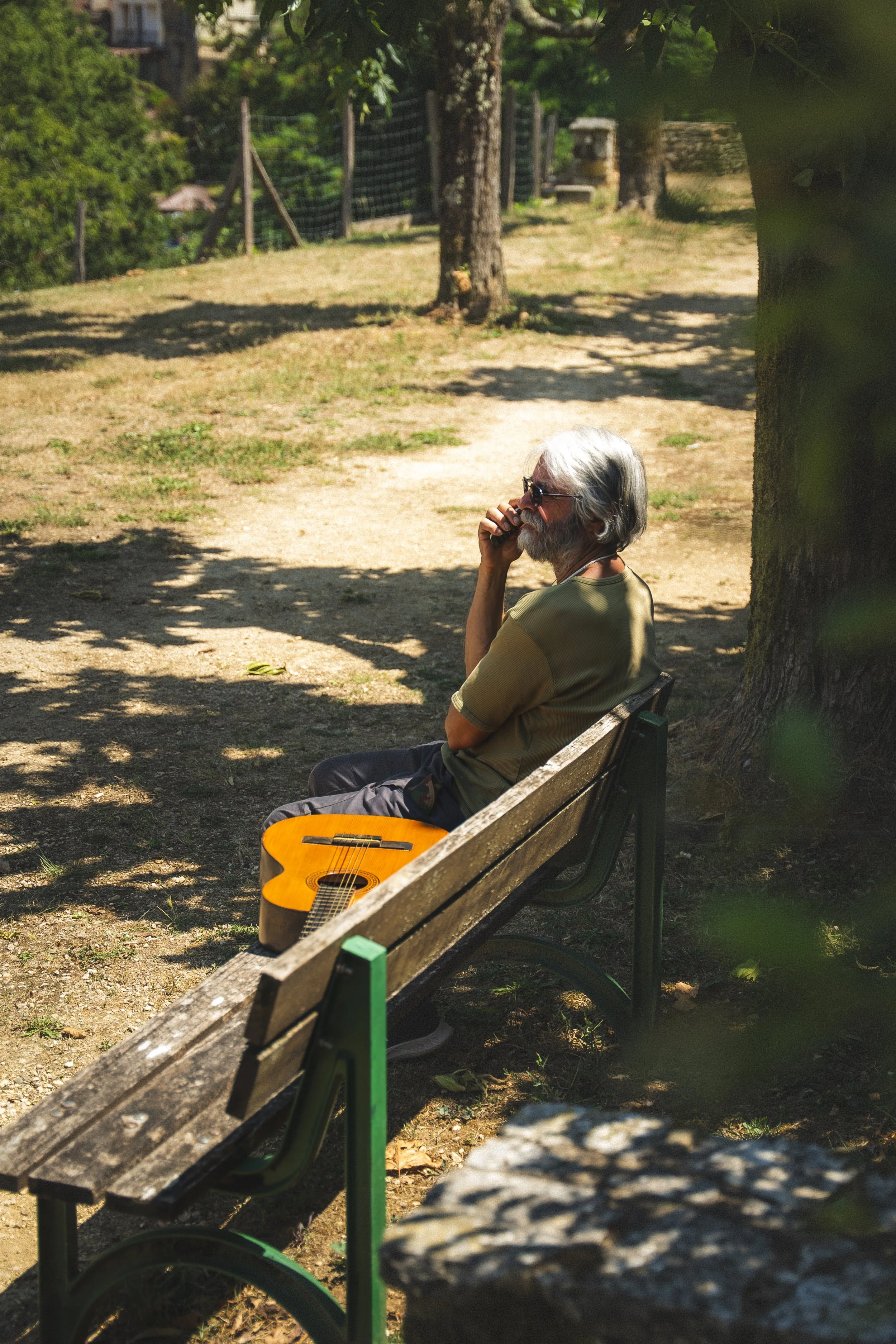Een oudere man zit op een parkbank onder een boom, met een gitaar op zijn schoot, terwijl hij aan het bellen is op een zonnige dag.