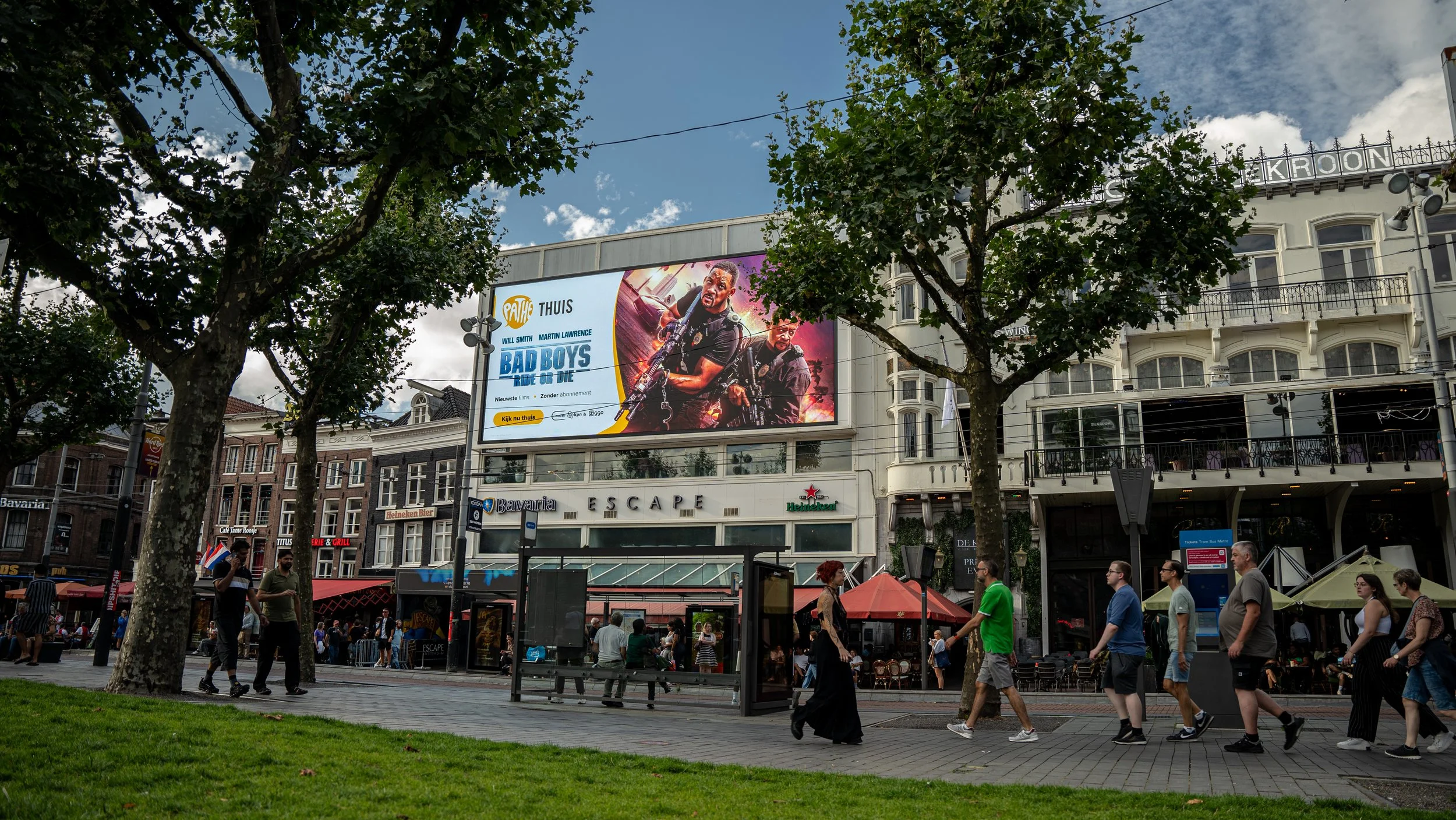 Straatzicht met mensen die lopen voor een winkelgebied met grote reclameborden en dichtbeboste bomen.