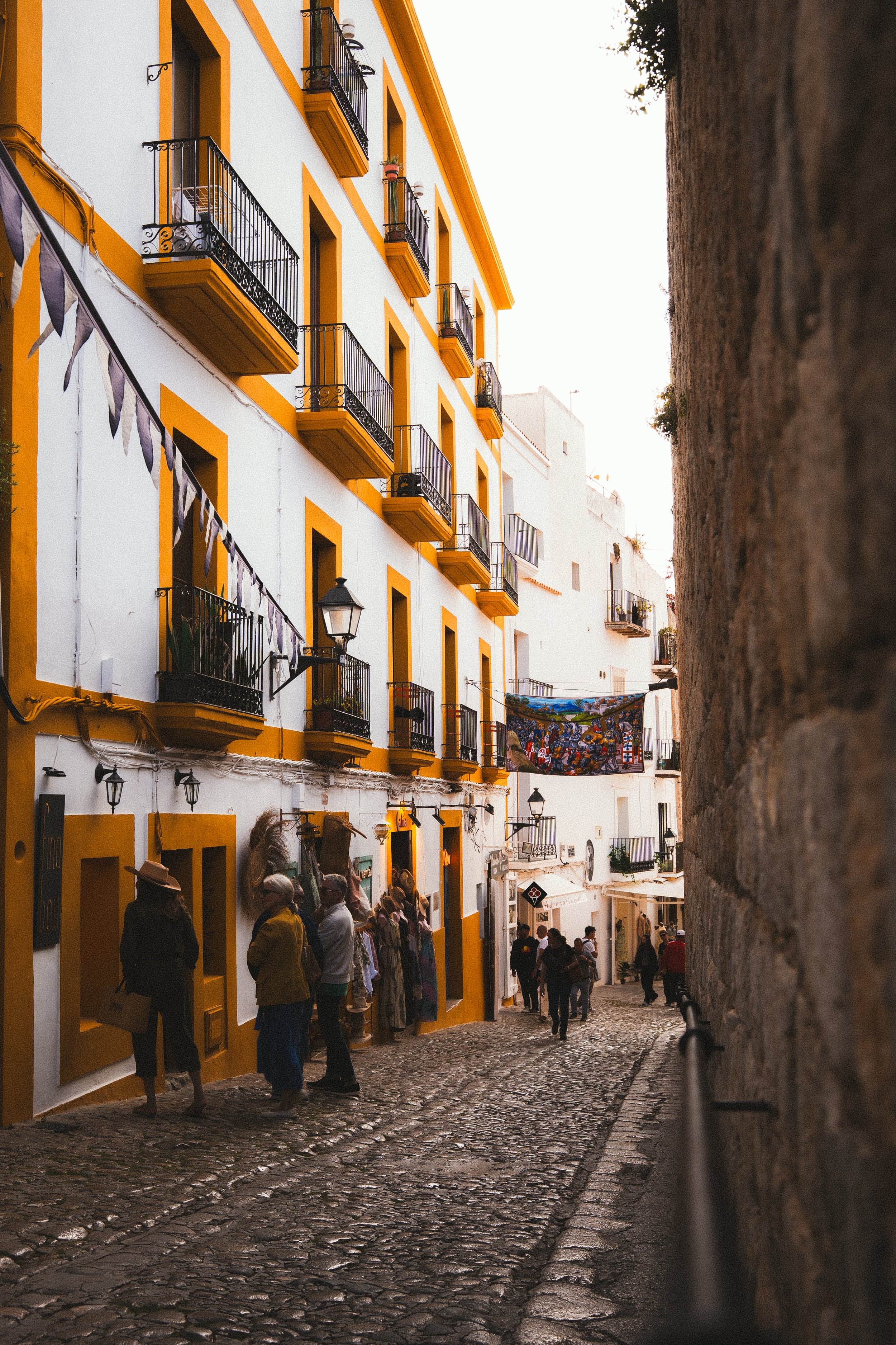 Een schilderachtige straat met witgepleisterde gebouwen met gele accenten en kleine balkons, terwijl mensen winkelen en door de straat lopen.