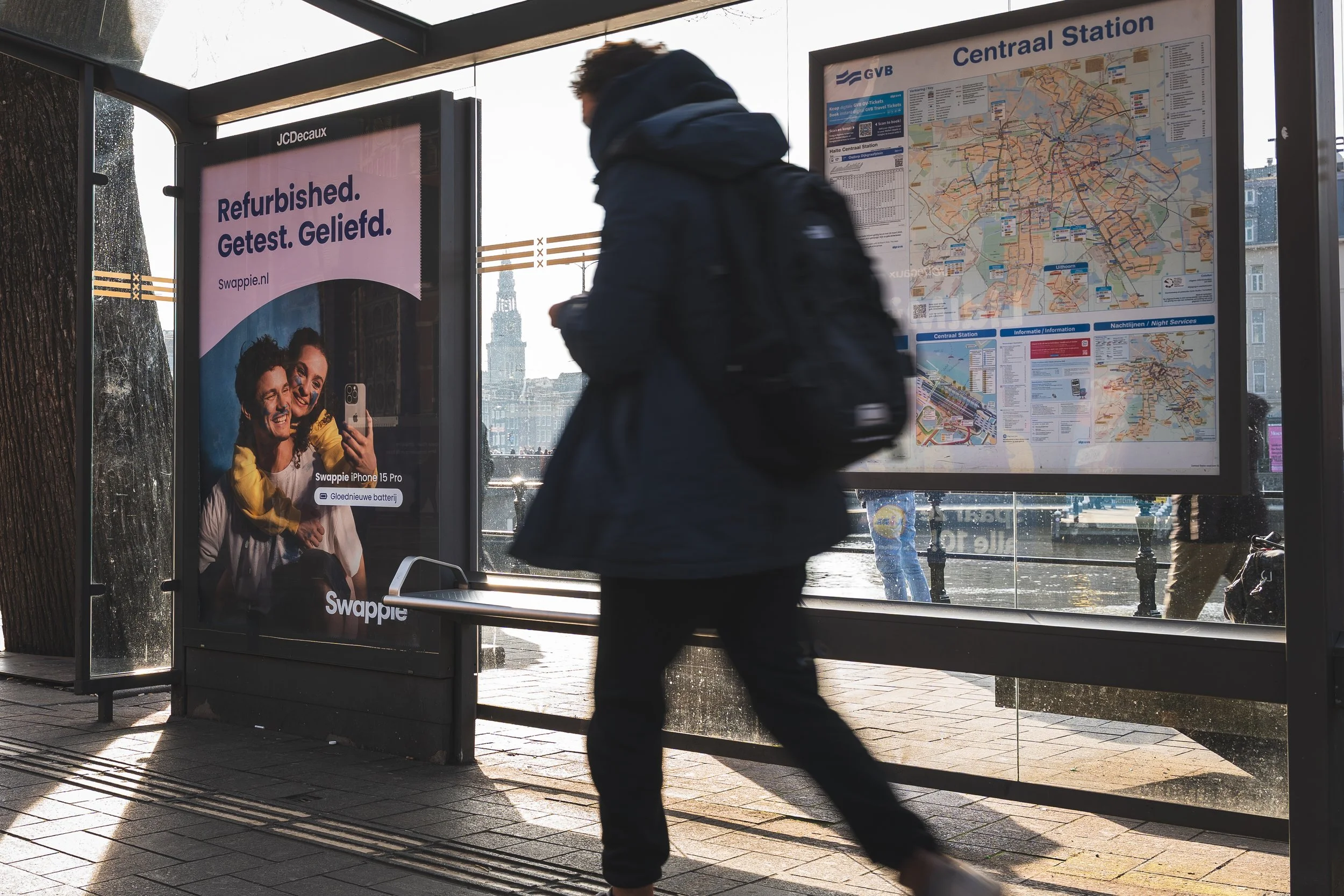 Een persoon met een blauwe jas en rugzak loopt langs een bushalte met een openbaar vervoersplan en een advertentie voor een iPhone 15 Pro, met een rivier en gebouwen op de achtergrond.