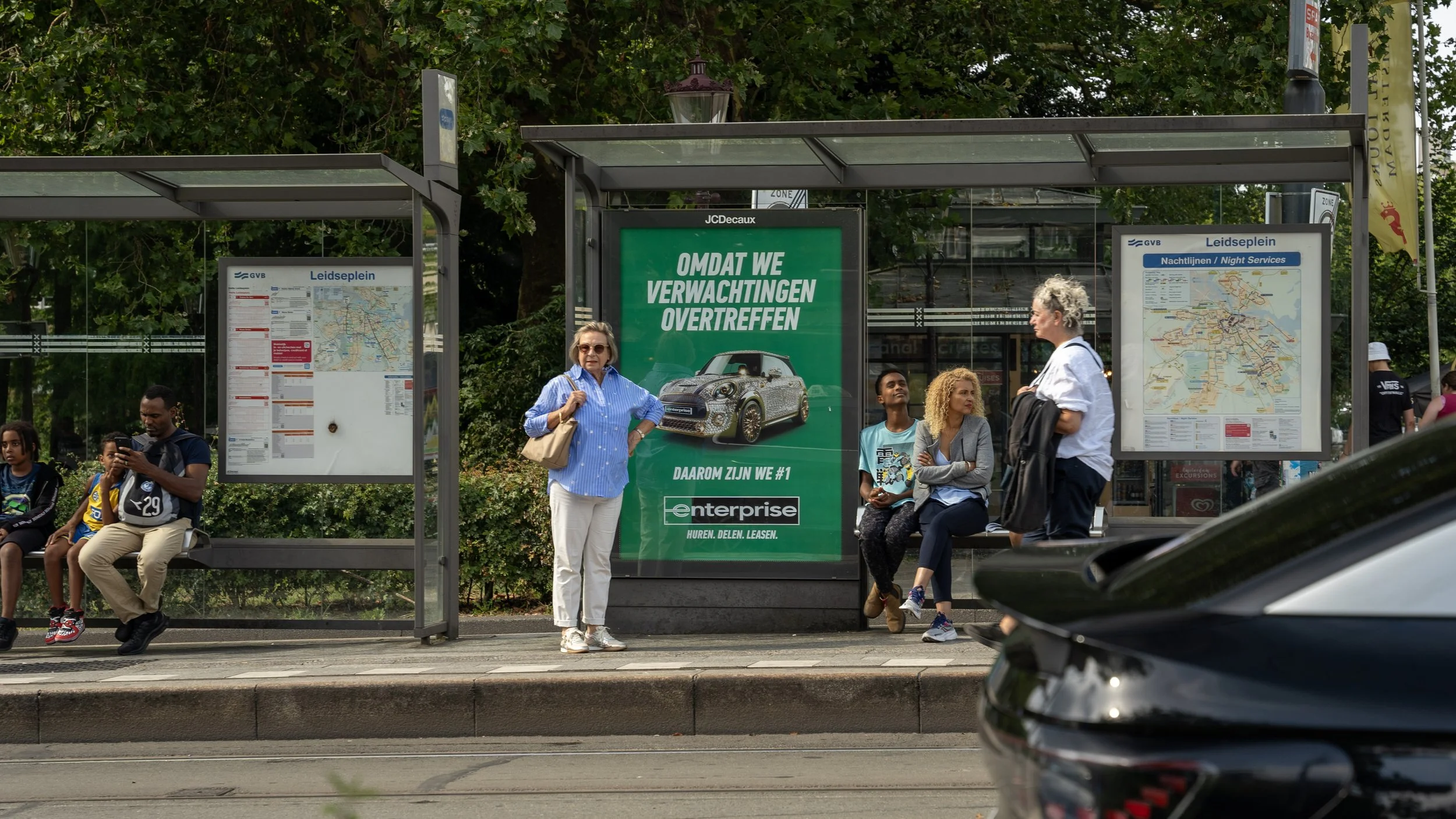 Grote groene reclameborden bij een bushaltes met mensen die wachten, waaronder een vrouw met witte haar en een vrouw met een grijze jas, en een jongen met een T-shirt. Op de achtergrond groene bomen en een deel van een auto.