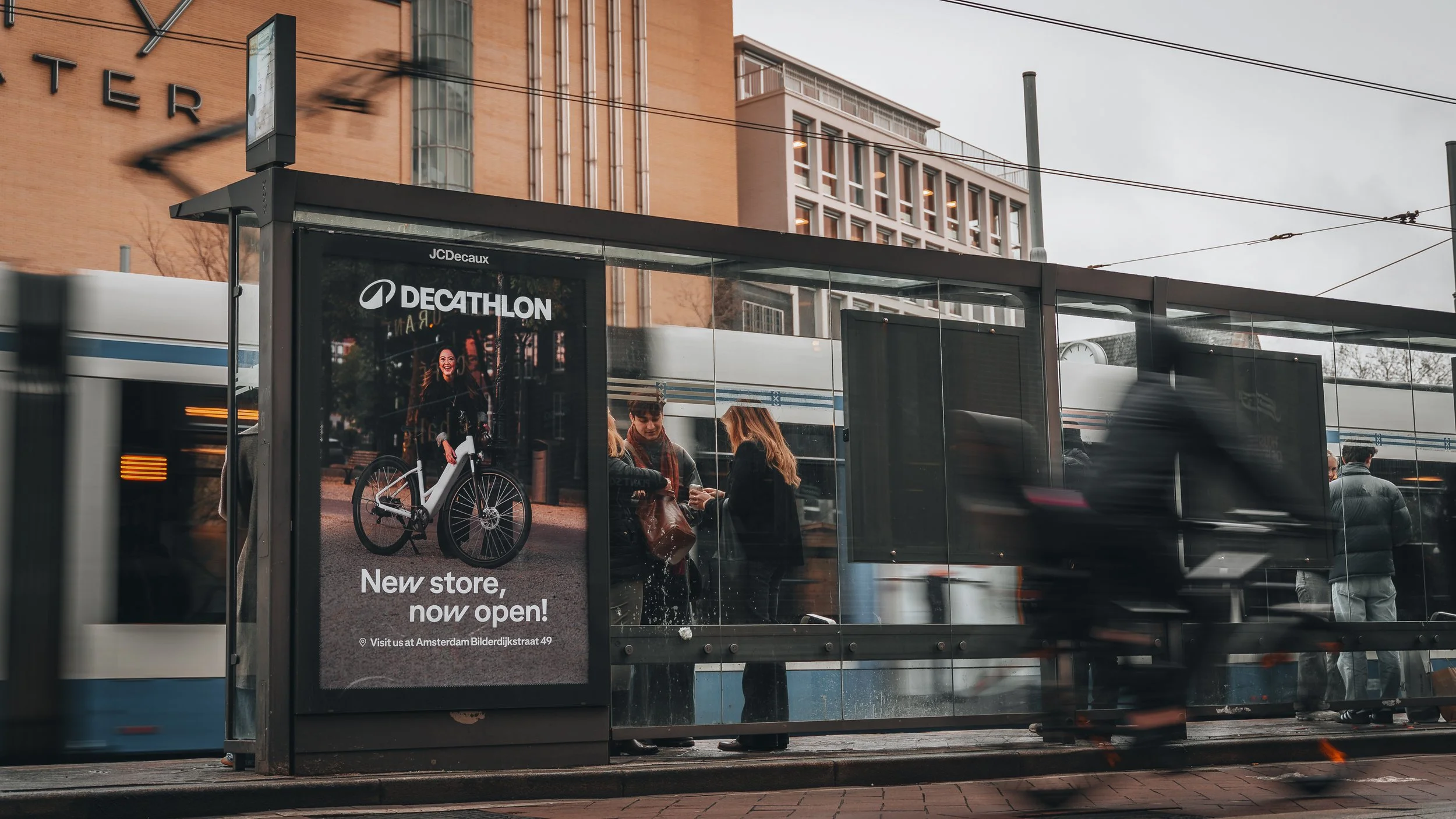 Mensen wachten bij een tramhalte, met een digitale advertentiebord dat een fiets en een vrouw toont, en een tram rijdt voorbij