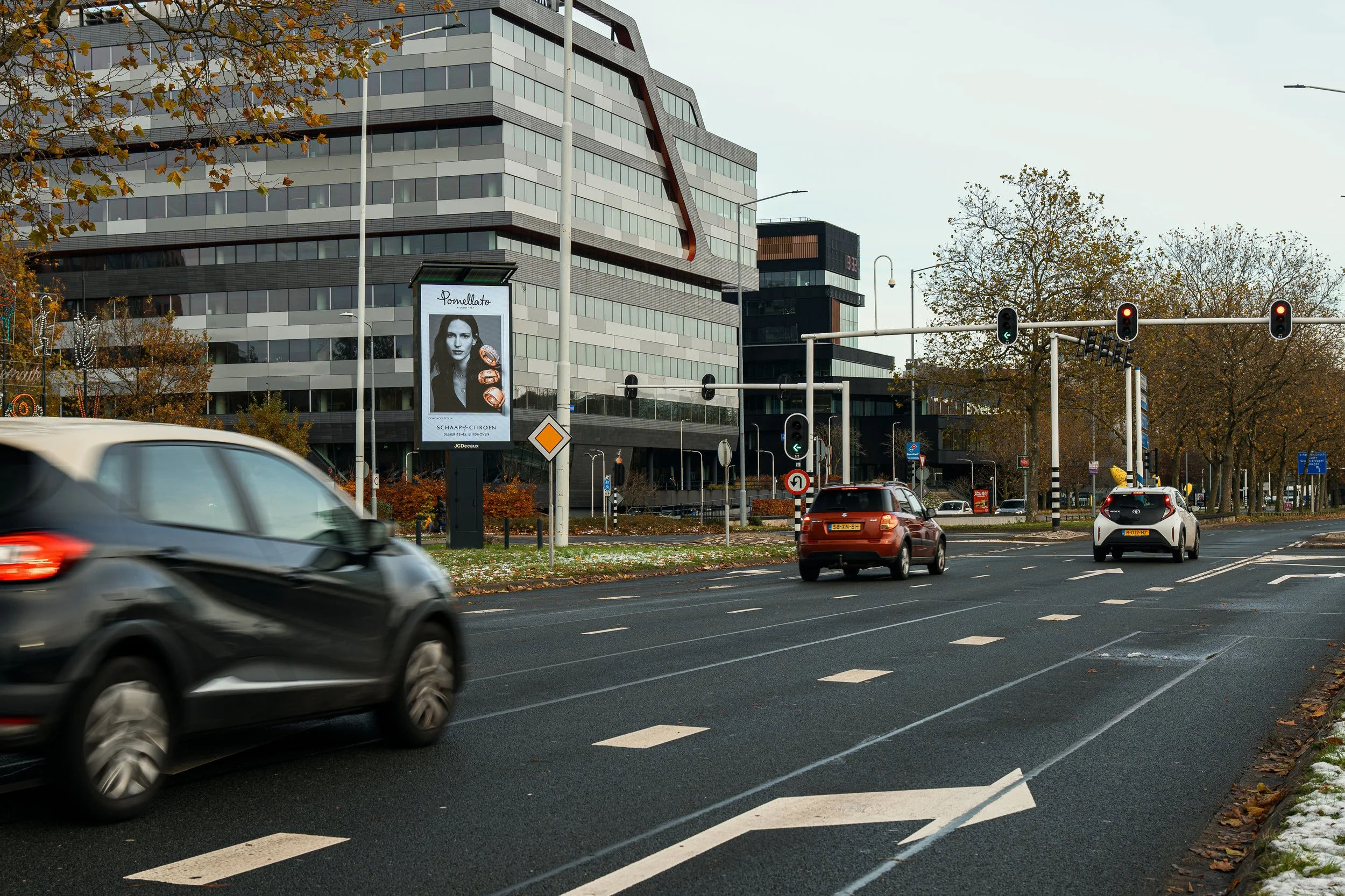 Straat met meerdere auto's die wachten op groen licht, verkeerslichten en een modern kantoorgebouw op de achtergrond, herfstbomen met herfstbladeren. 