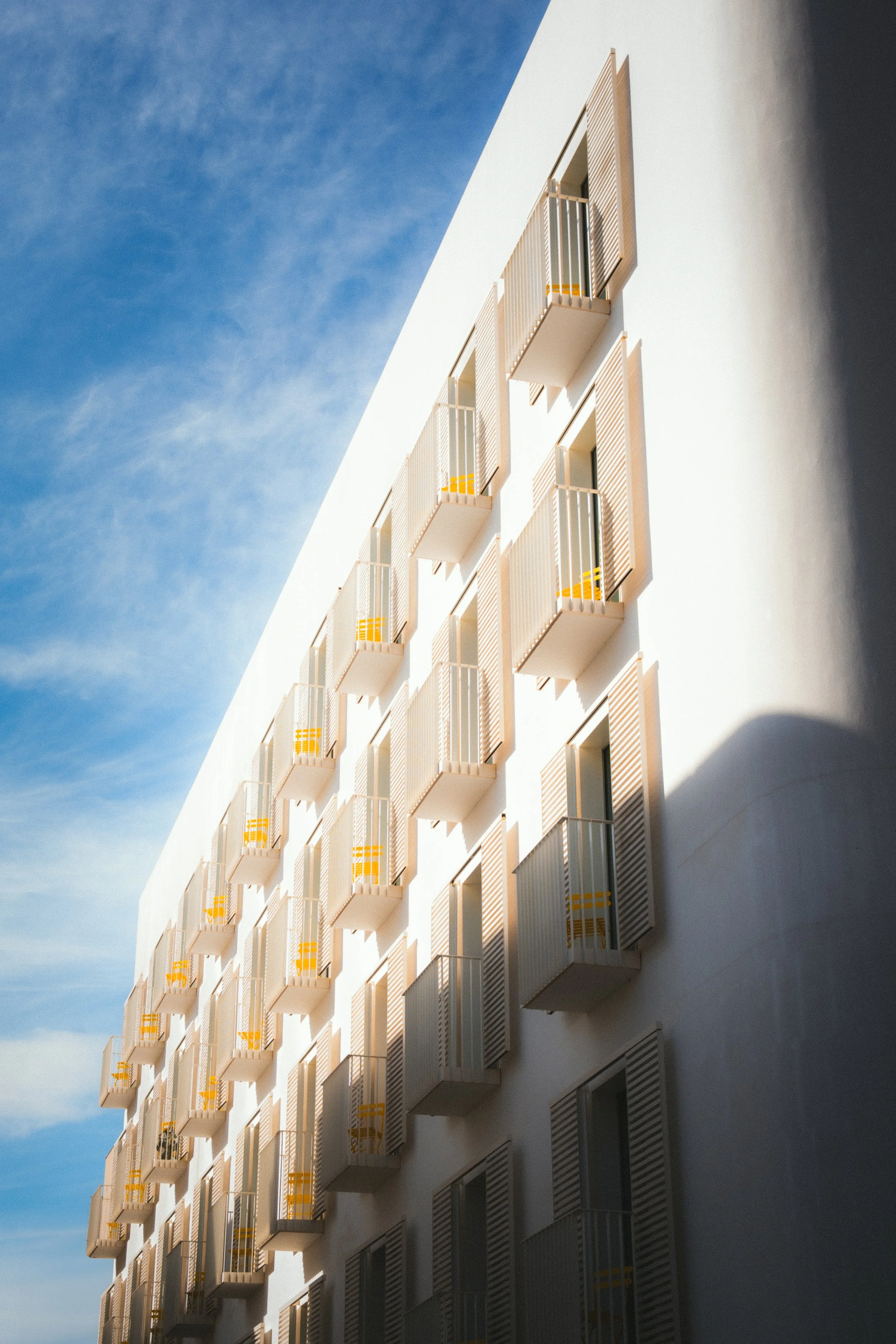 Modern wit appartementengebouw met kleine balkons en open luiken, onder een blauwe lucht.