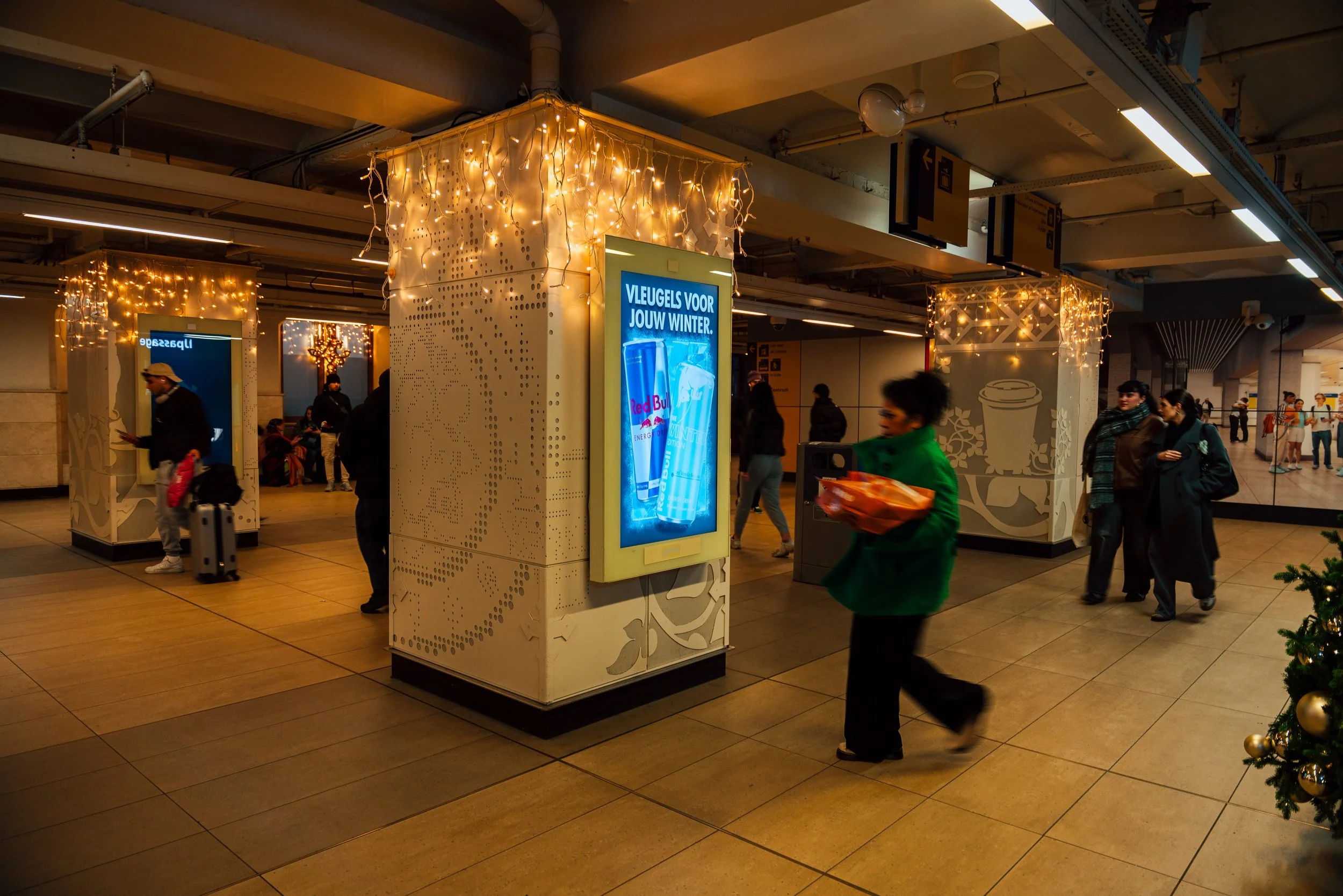 Binnen een winkelcentrum of station met decoraties en mensen die wachten of doorlopen. Decoraties bestaan uit lichte stringverlichting en witte panelen met kerst- of winterthema.