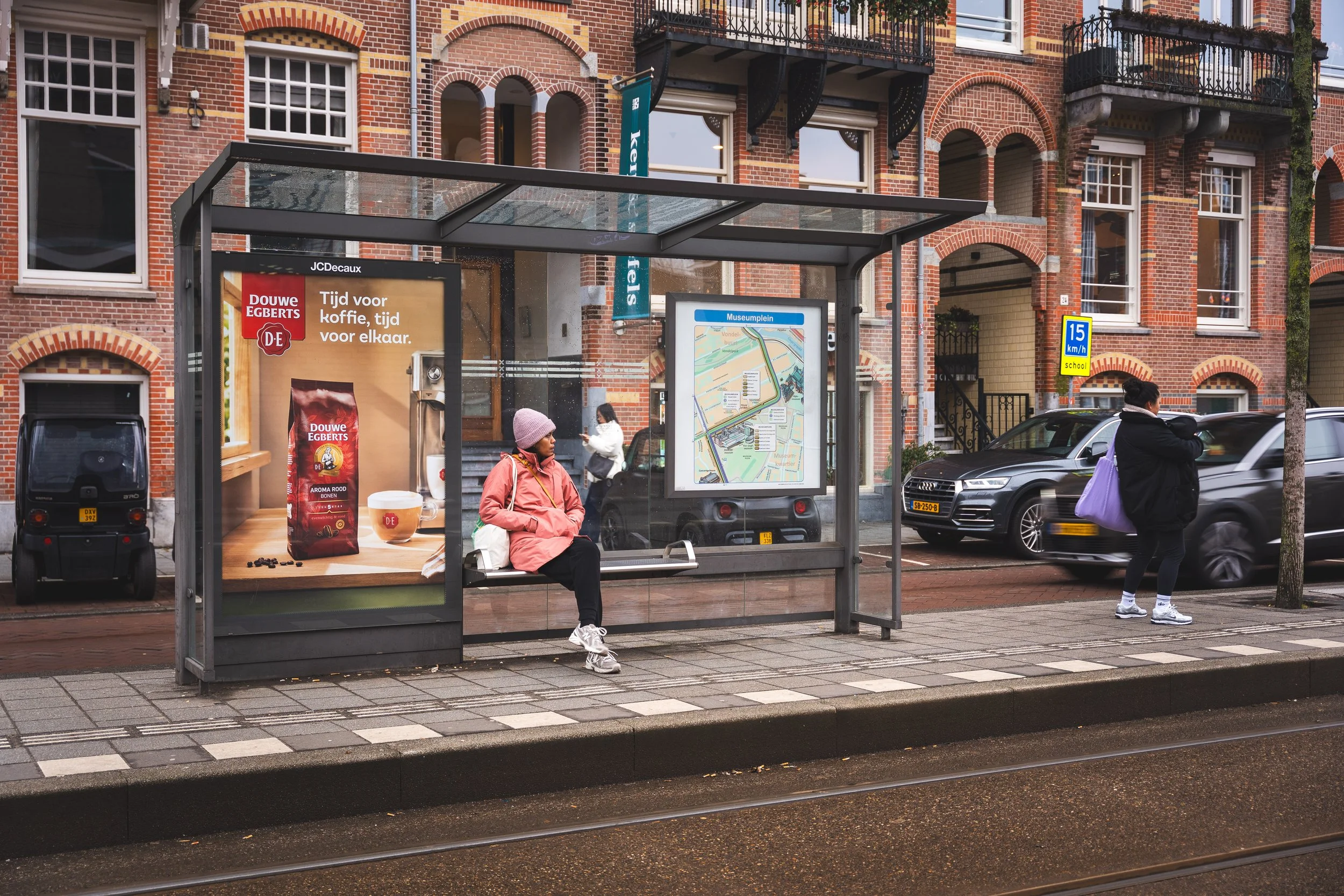 Een bushalte met een pasagier die op een bankje zit, een persoon die iemand anders belt, en een reclamebord voor Douwe Egberts koffie, met een stadskaart en gebouwachtergrond.