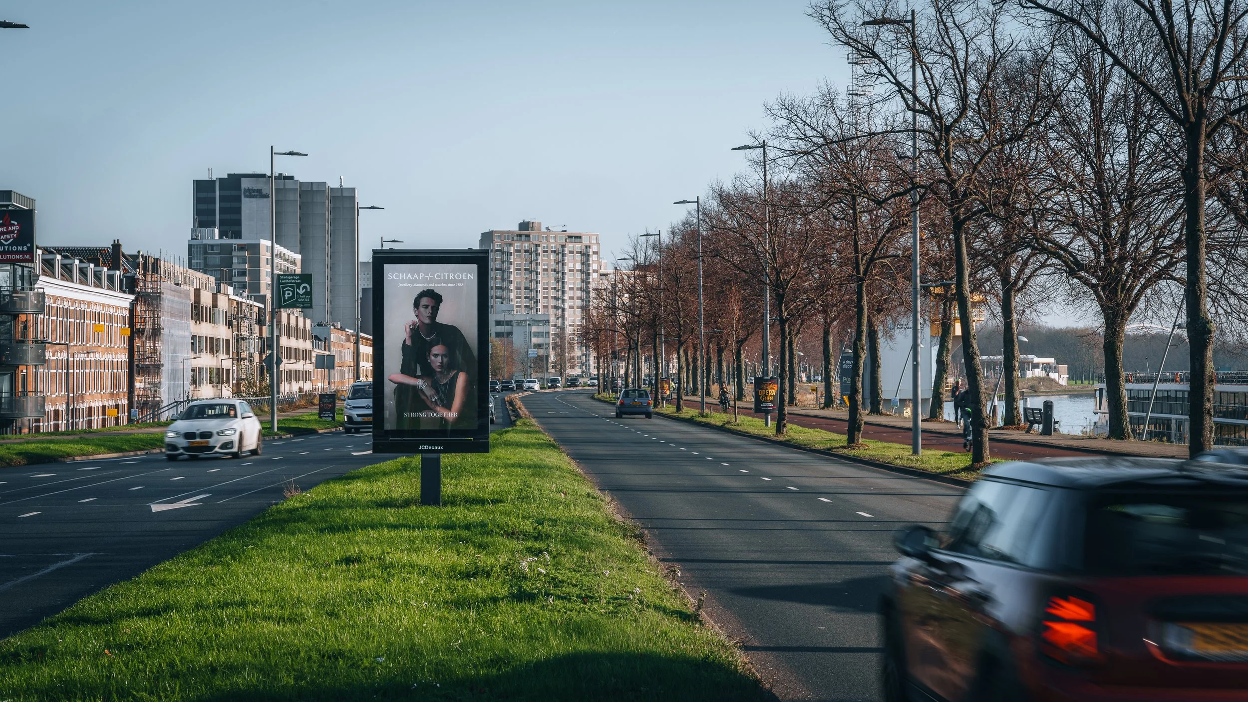 Een straat met auto's en een billboard met fashionfotografie. Er zijn bomen langs de weg en gebouwen op de achtergrond, onder een blauwe hemel.