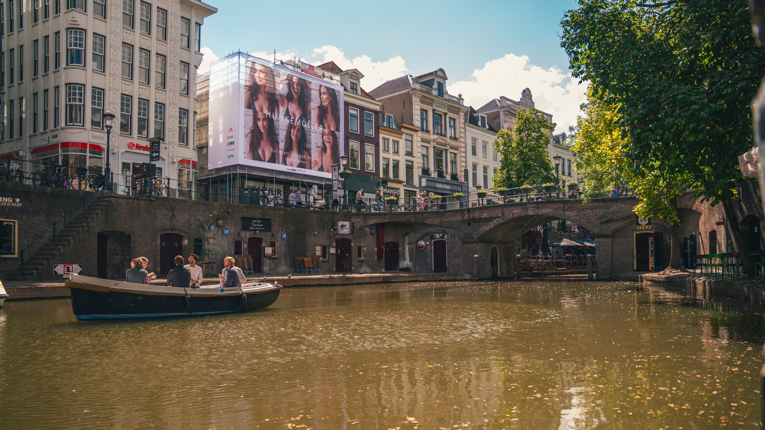 Een grachten in Amsterdam met een kleine boot en vier mensen erin. In de achtergrond hangen historische gebouwen en een groot reclamebord met een model van haar.
