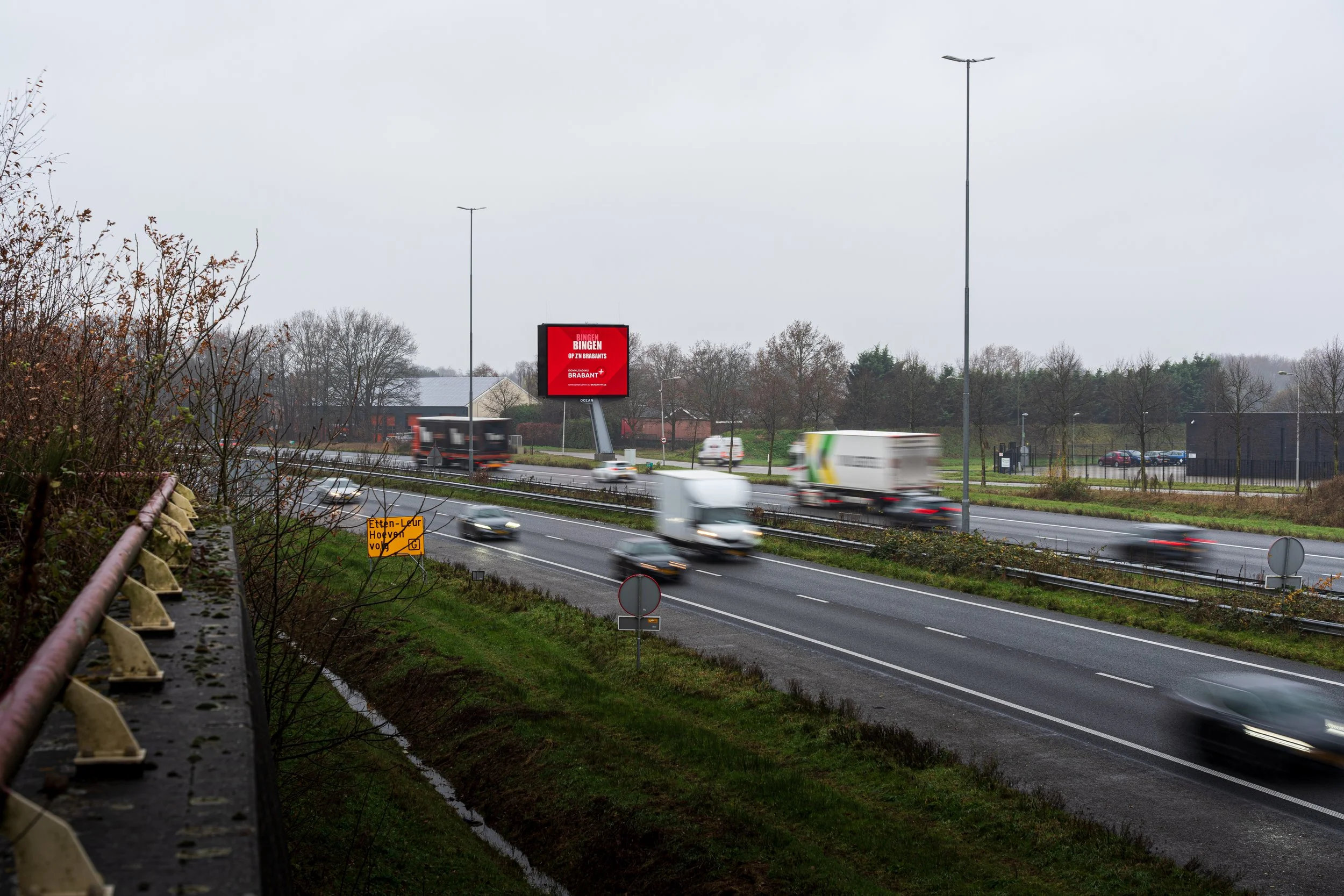 Snelweg met verkeer en bewegende auto's op een grijze dag, bomen en een billboard op de achtergrond.