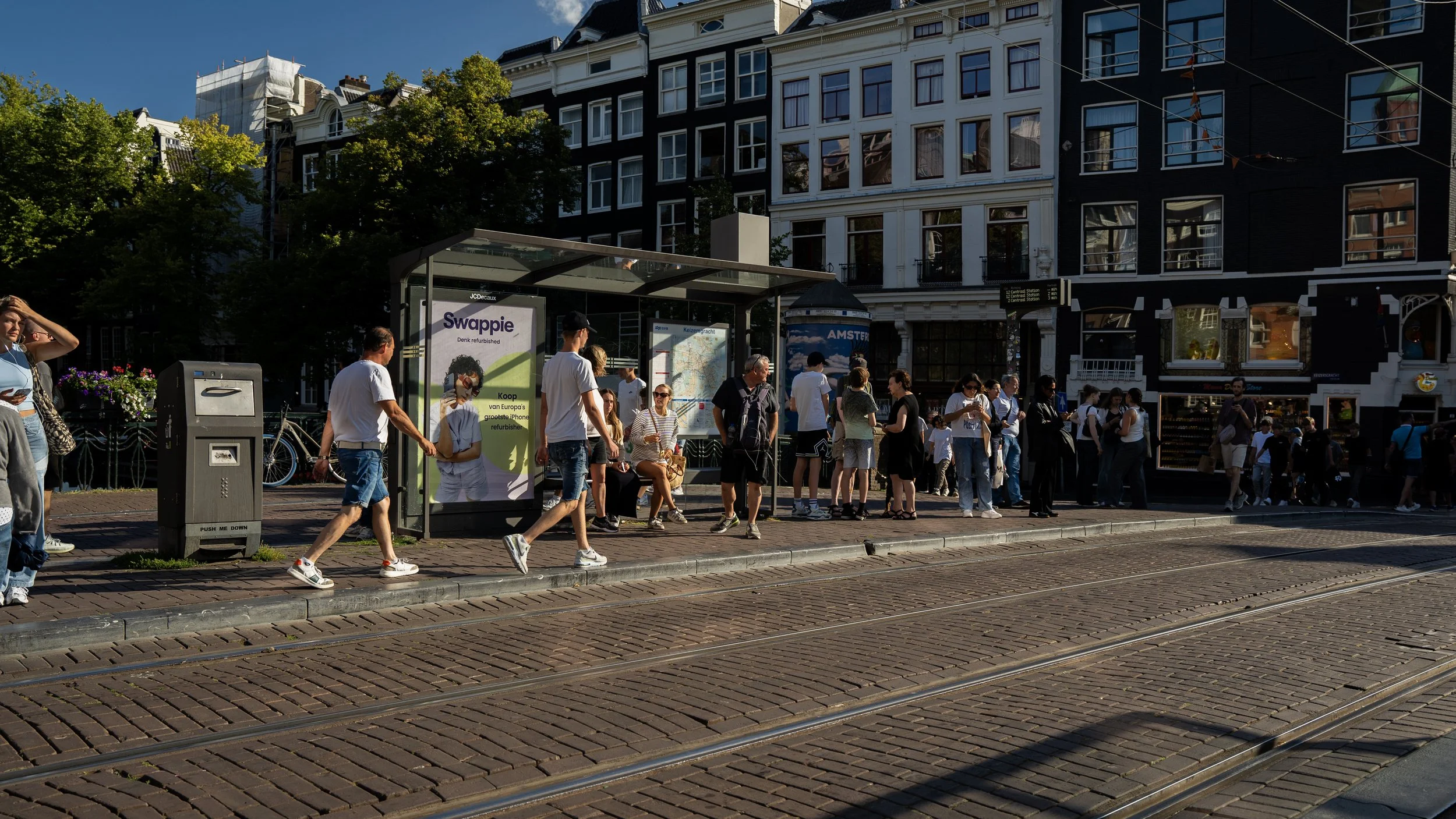 Mensen wachten bij een bushalte op een zonnige dag in een stad met traditionele gevels en tramrails op de weg.
