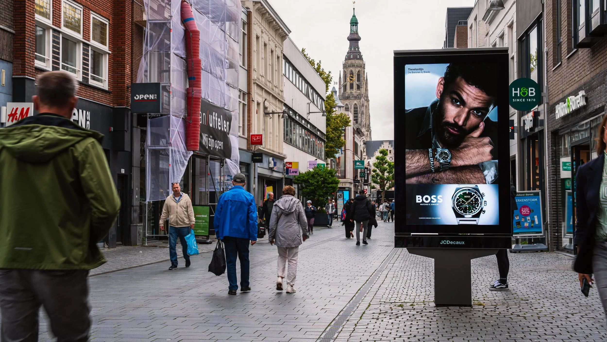 Een drukke winkelstraat met mensen die lopen en winkeltjes aan weerszijden. Een groot digitaal reclamebord toont een man die een horloge draagt, met een tekst voor het horlogemerk BOSS.