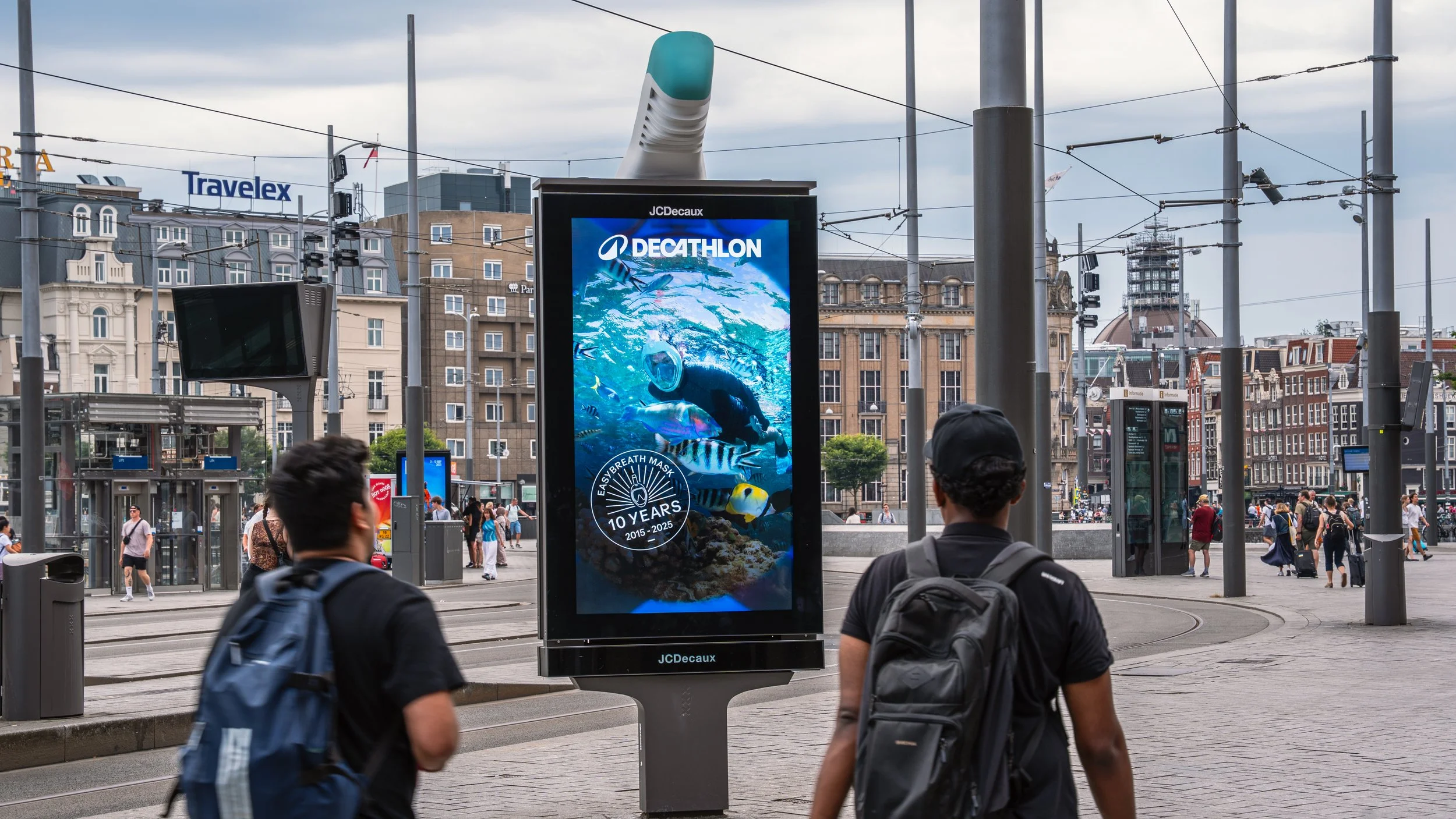 Het image toont een stadsplein met een elektronisch reclamebord dat een duiker met onderwaterdieren weergeeft en een afbeelding van een grote hand die eruitziet als een opblaasbare duim. Er lopen meerdere mensen met backpacks en er zijn gebouwen en t
