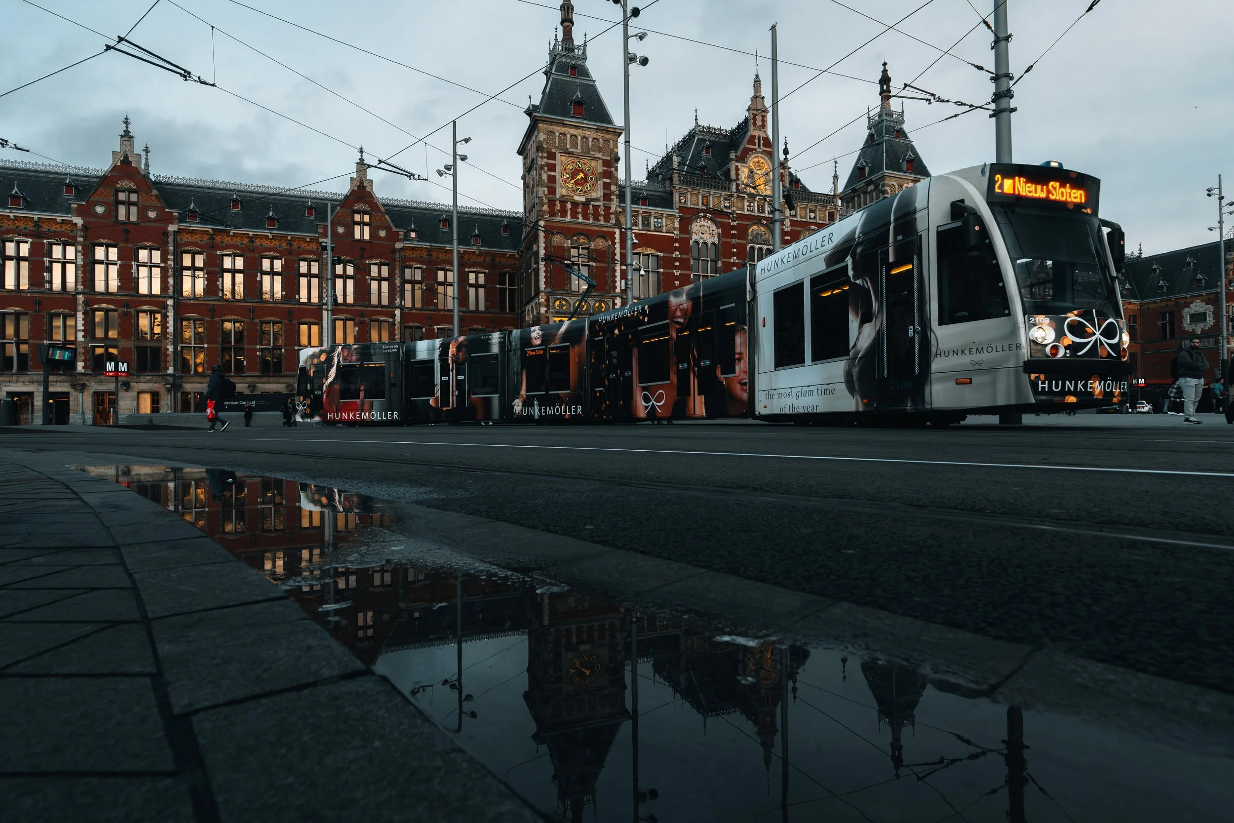 Een tram met reclame in een historische stadssituatie, vlakbij een oud stationgebouw met torens en een klok, onder een bewolkte hemel.