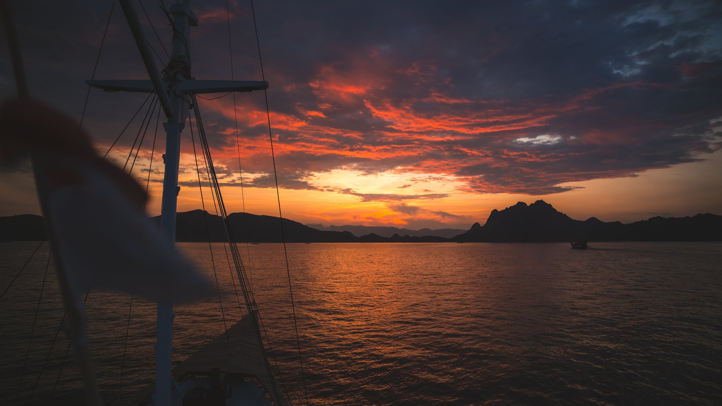 Uitzicht over de zee bij zonsondergang met een silhouet van een zeilboot en bergen op de achtergrond, met oranje en paarse kleuren in de lucht.