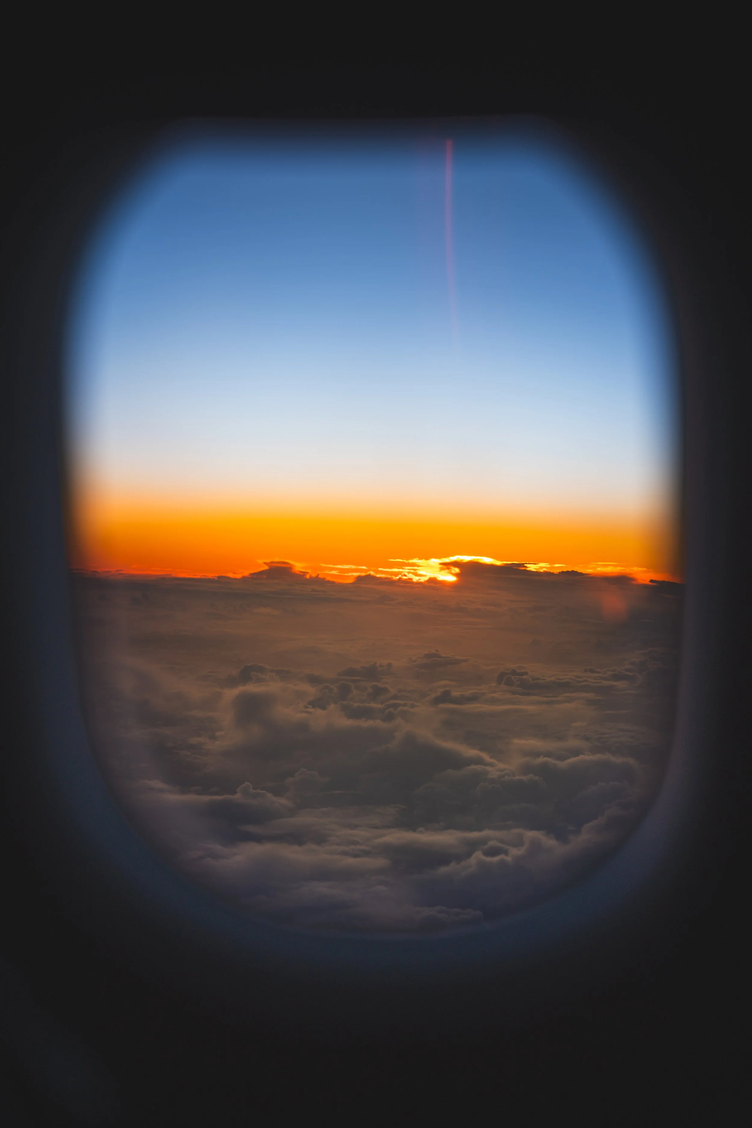 Luchtfoto van een zonsondergang genomen vanuit een vliegtuigraampje met wolken onder en een heldere blauwe lucht boven.