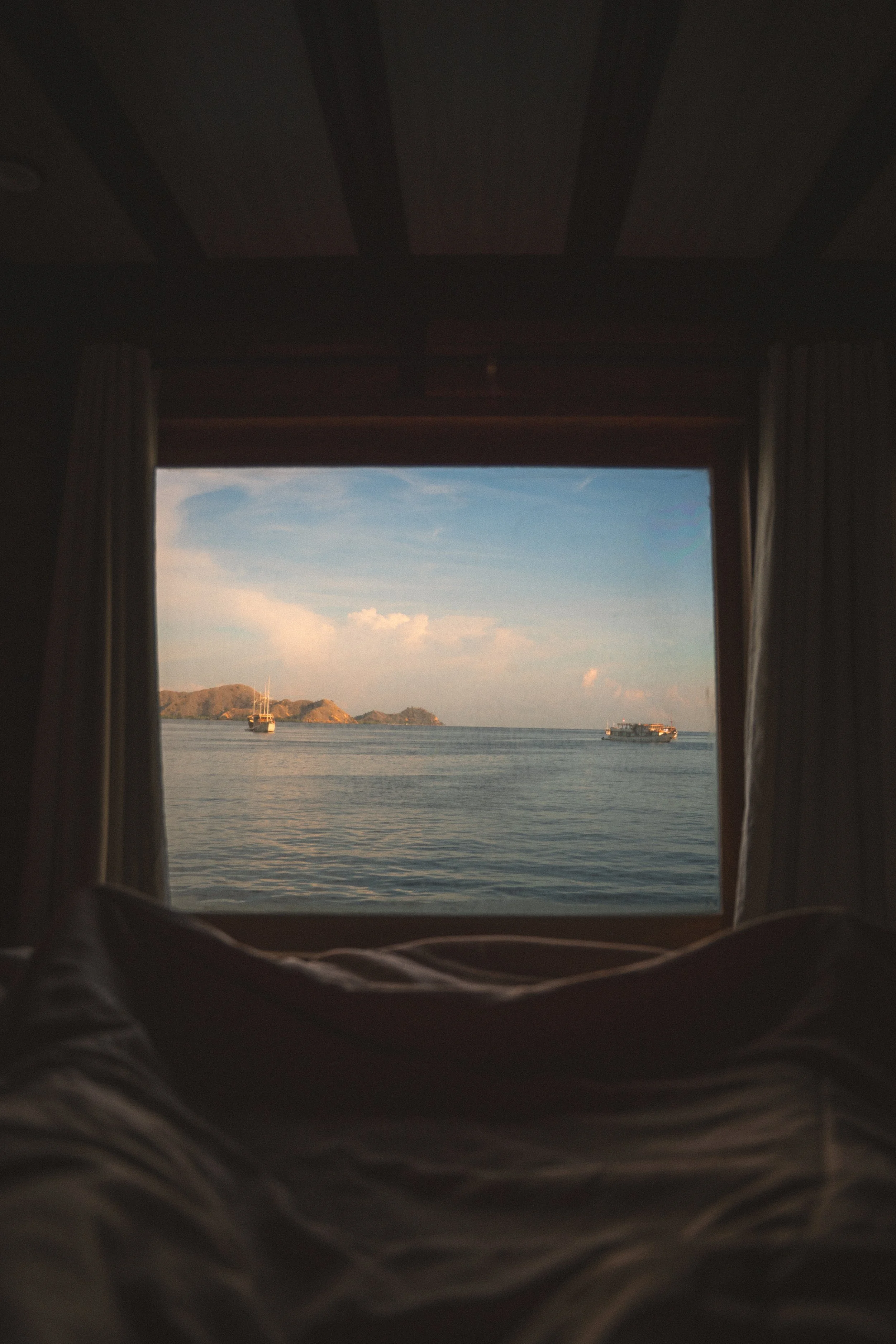 Uitzicht vanaf een kamer met uitzicht op de zee, met boten en kleine eilanden in de verte, gezien door een raam met gordijnen.