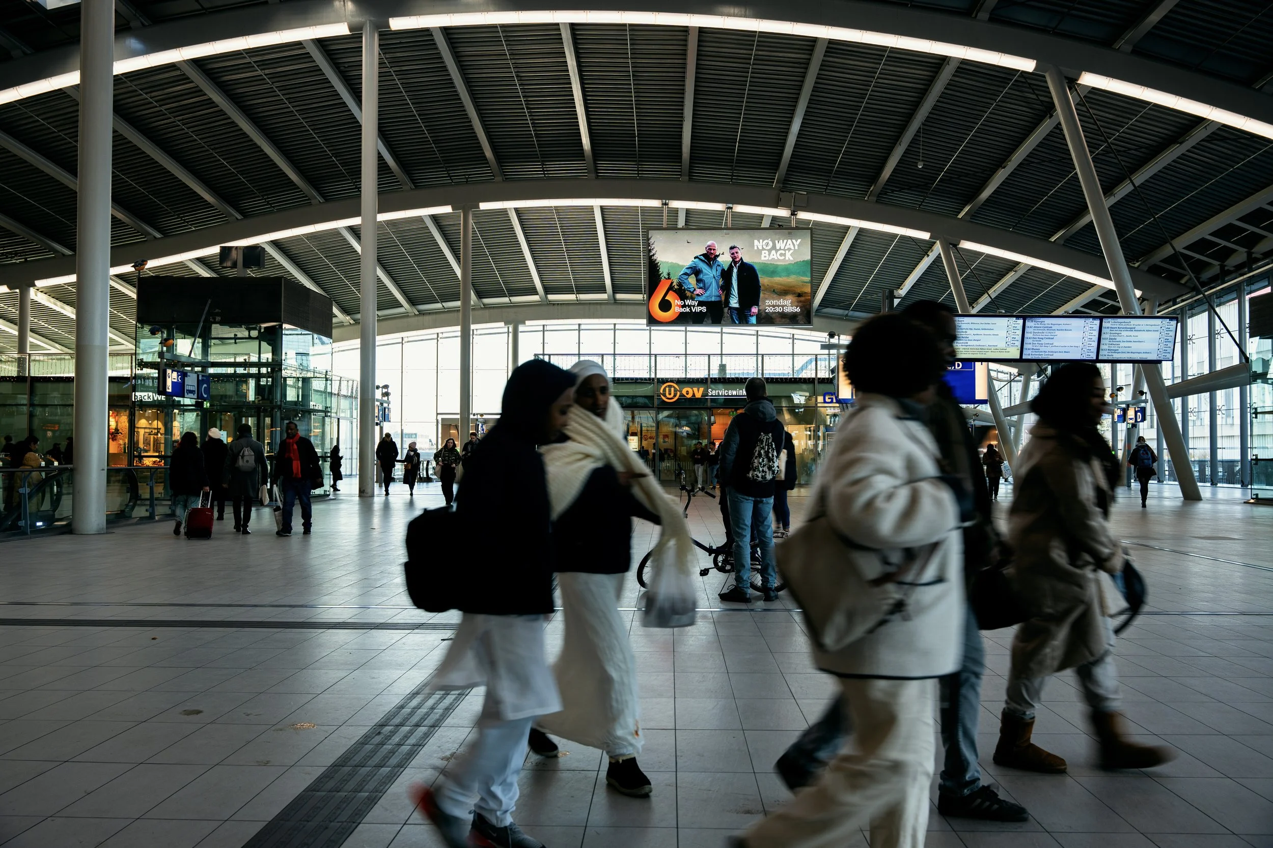 Binnenkant van een druk treinstation met mensen die lopen, check-in balies en digitale vertreklijsten.
