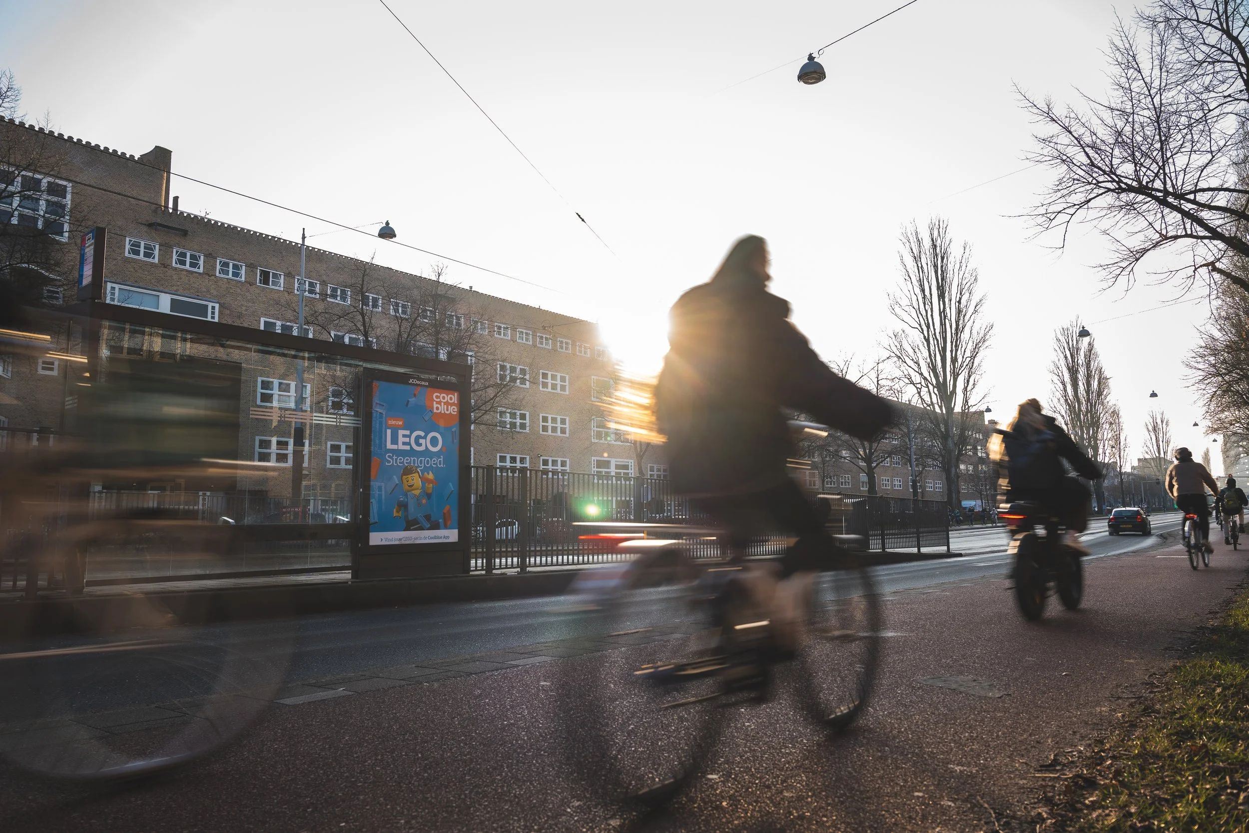 Mensen fietsen langs een straat met bomen en een gebouw, met de zon op de achtergrond en een billboard dat reclame maakt voor LEGO.