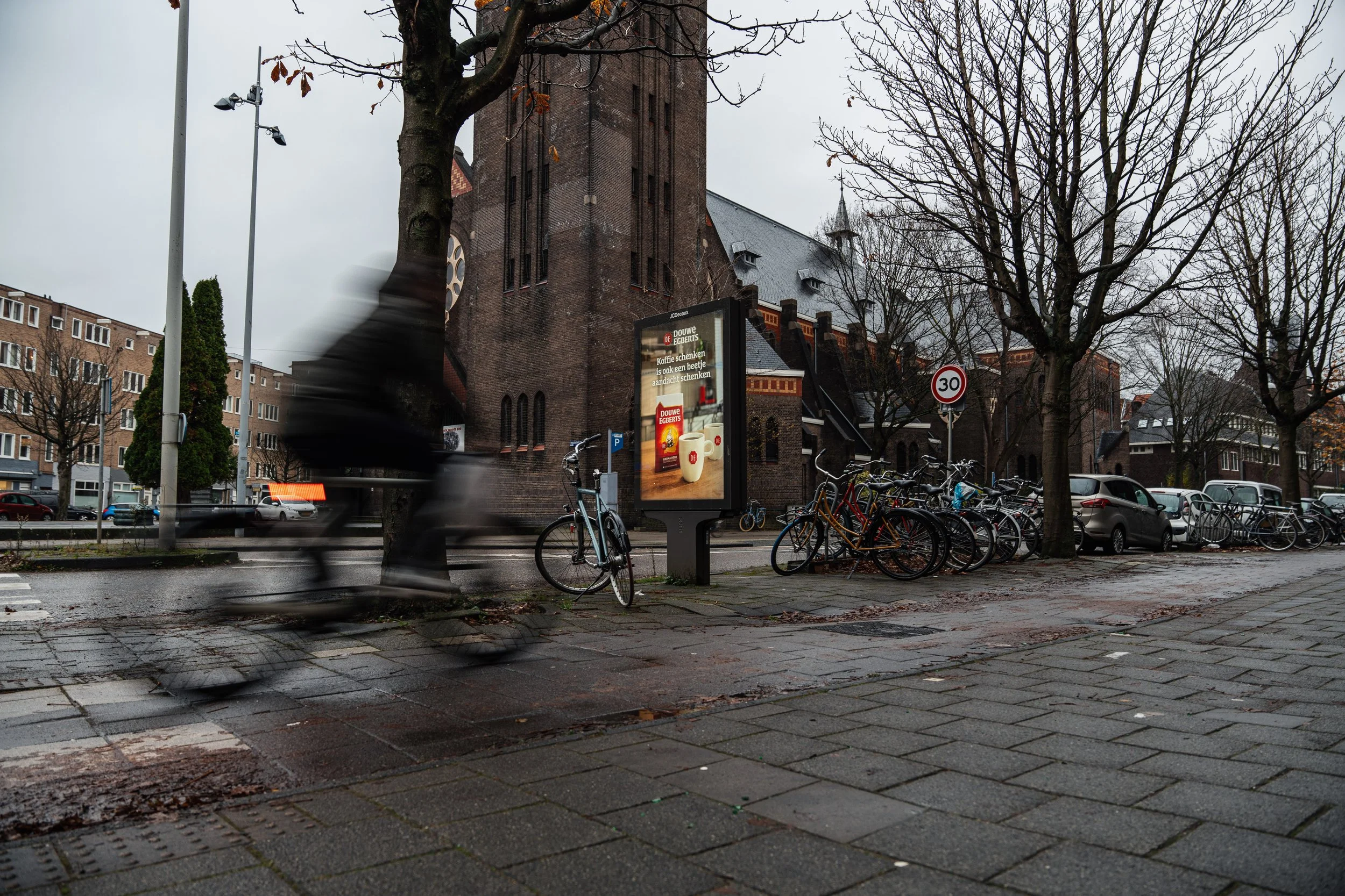 Een straat met een gebarsten trottoir, meerdere fietsen geparkeerd, een fietser die snel langs rijdt, een kerk met een toren, bomen zonder bladeren en een verkeerbord met limiet van 30 km/u.