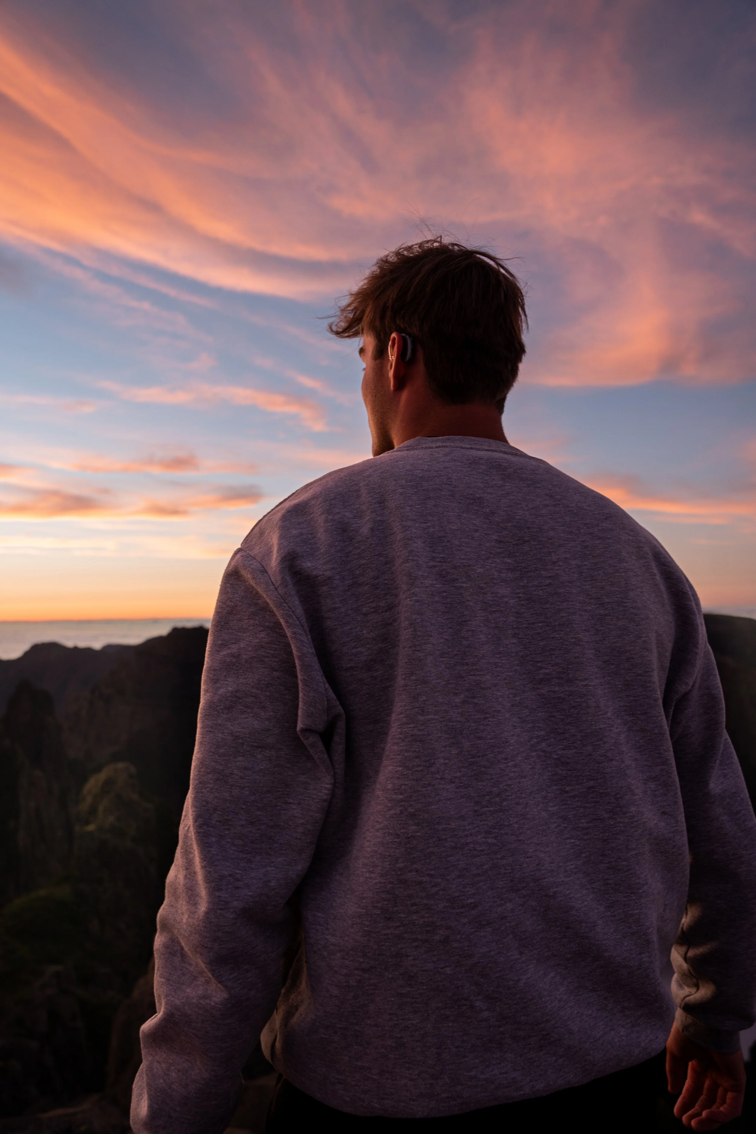 Man staat op een rots, kijkt naar de zonsondergang en draagt een gray sweatshirt en een oortje.