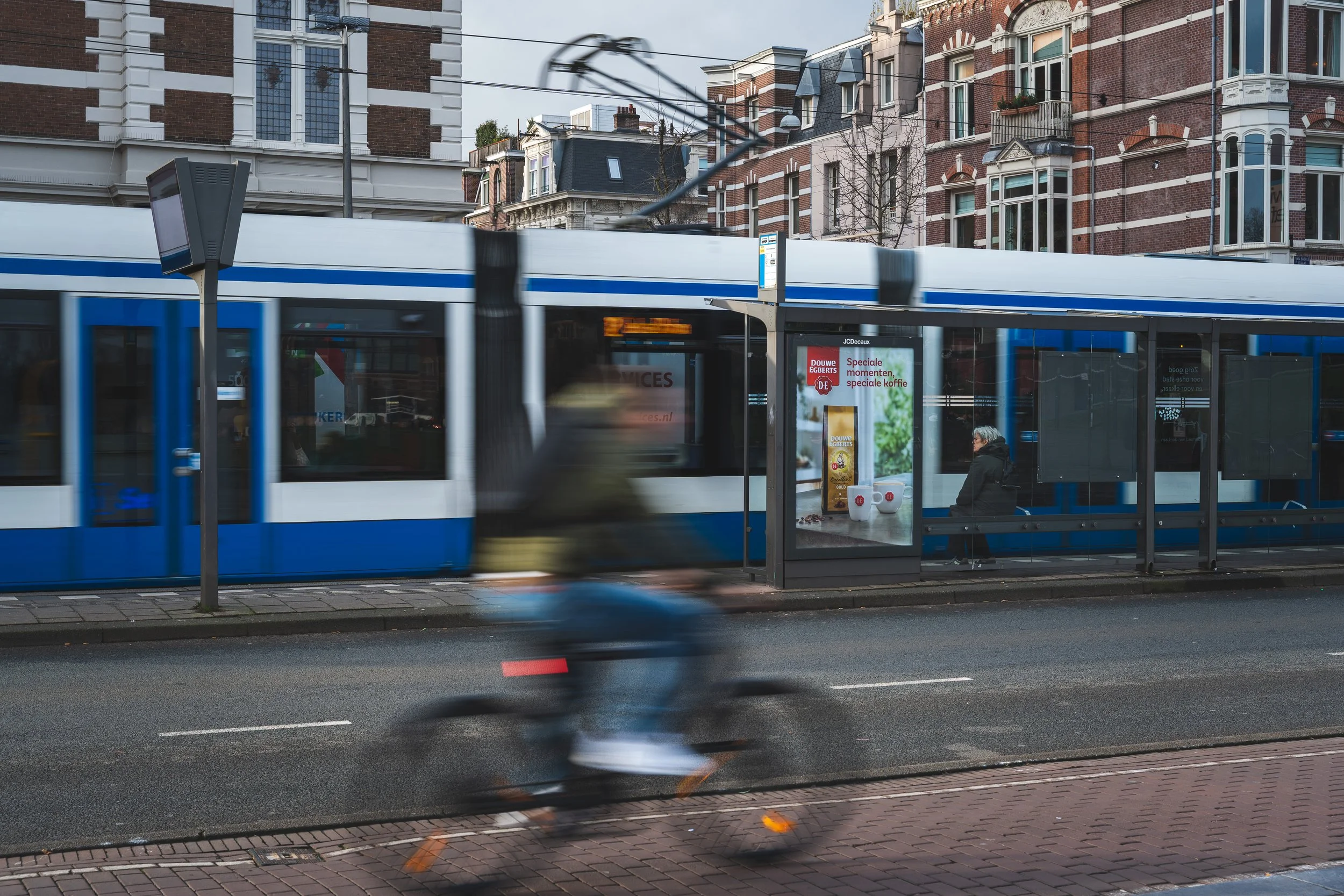 Een tram rijdt voorbij een bushokje waar een oudere persoon zit te wachten, terwijl een fietser snel passeert op de straat in een stedelijk gebied met historische gebouwen.