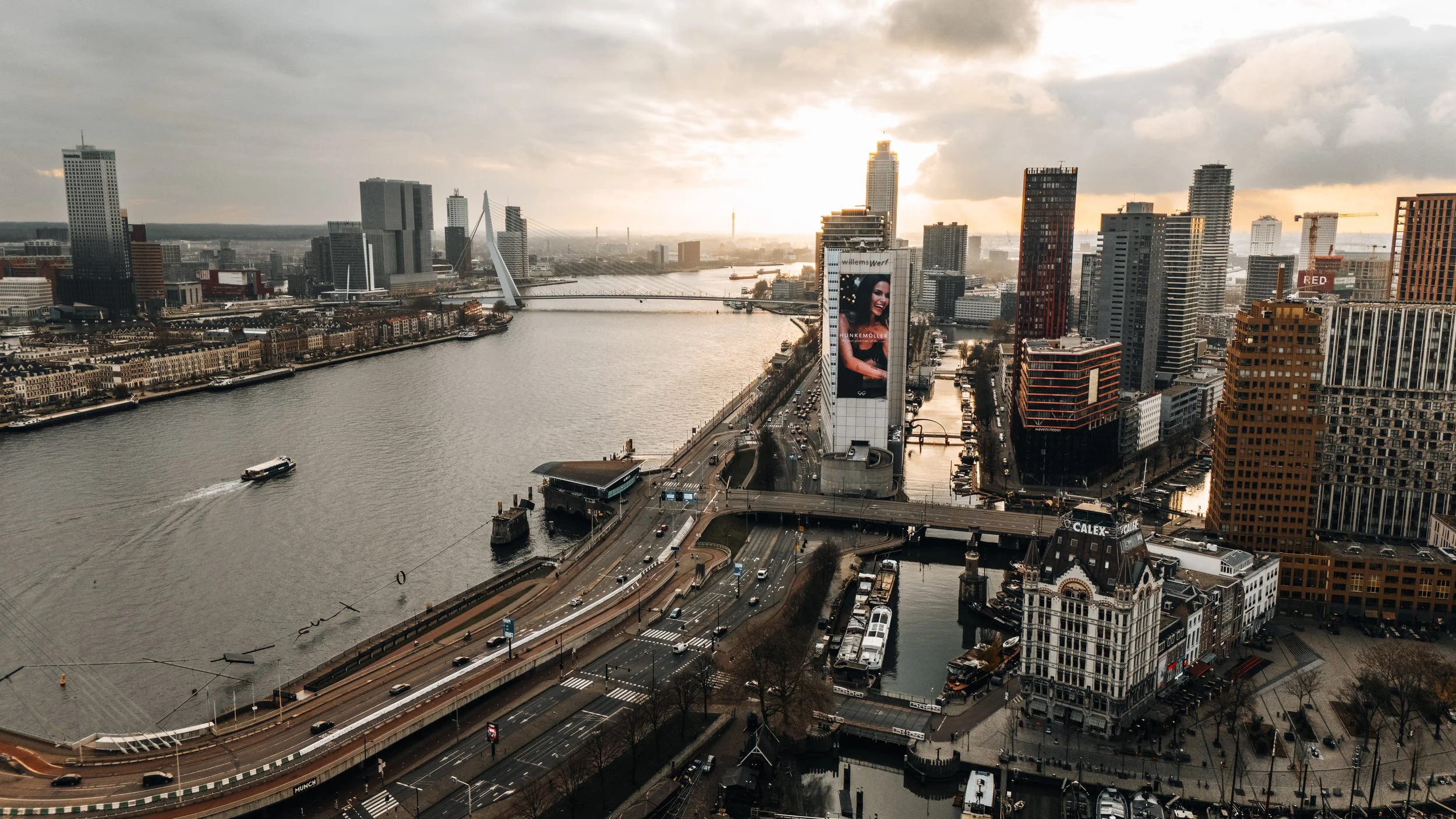 Stedelijke skyline met hoge gebouwen langs rivier en bruggen bij zonsondergang in Rotterdam.