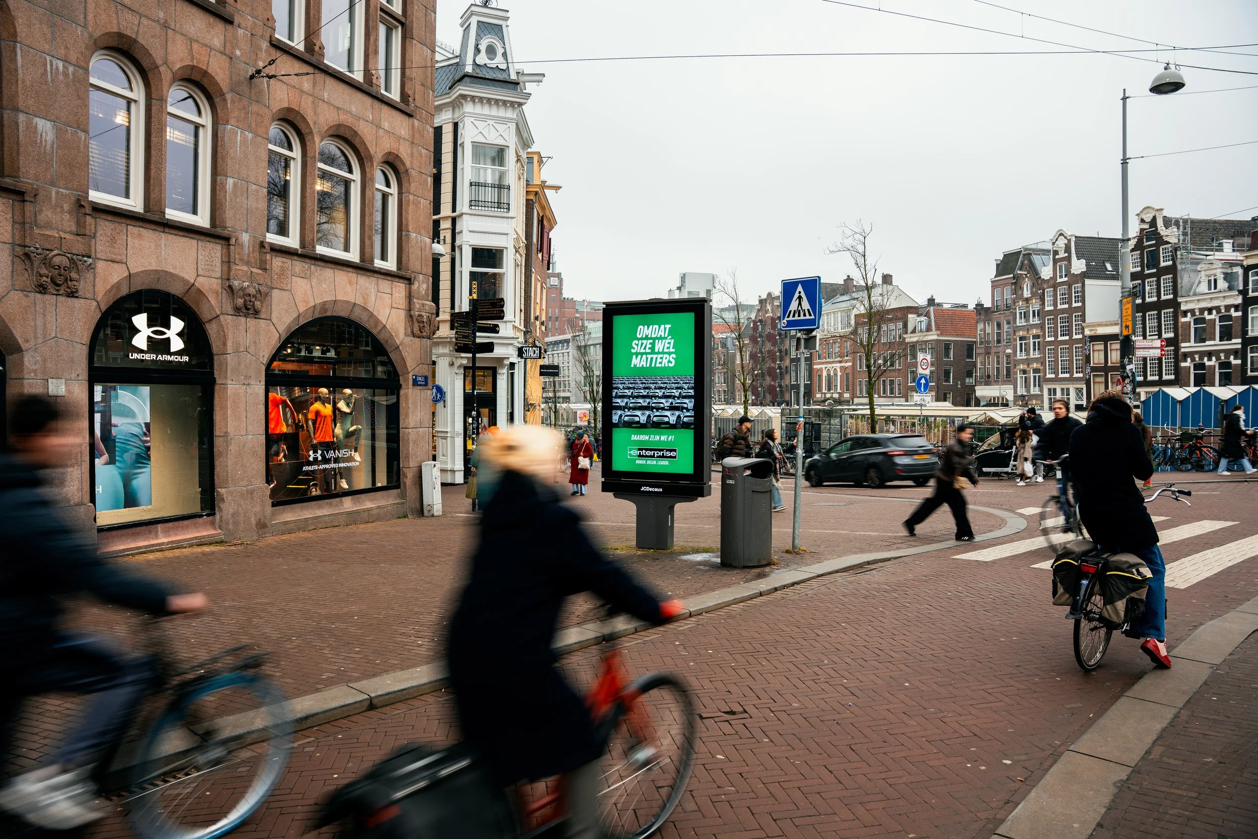Een druk straatbeeld met fietsers, voetgangers en een digitale reclamezuil in een stedelijke omgeving.