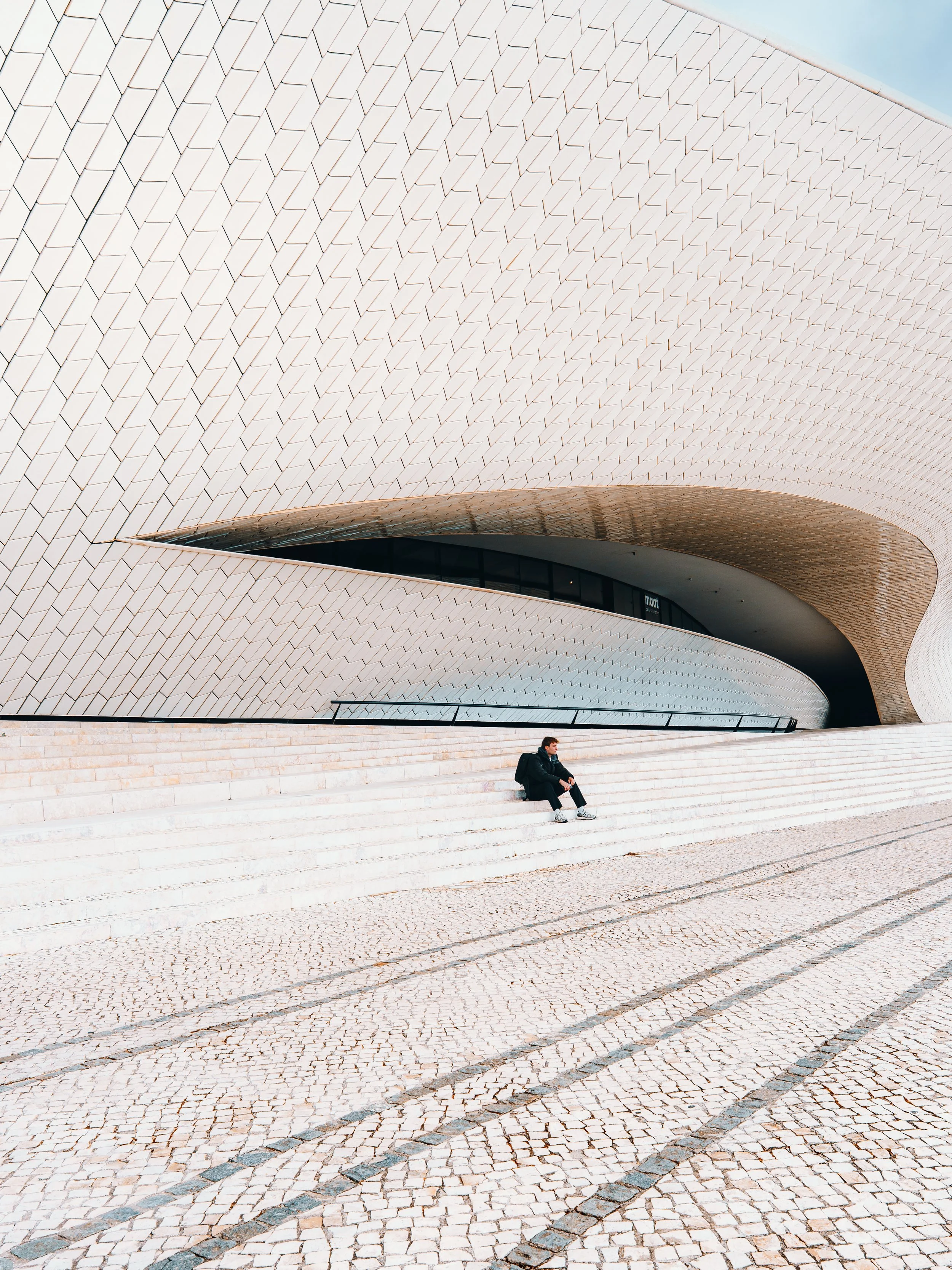 Een persoon zit op trappen buiten een modern, architectonisch gebouw met gewelfde muren en een geometrisch patroon op de gevel, onder een heldere hemel.